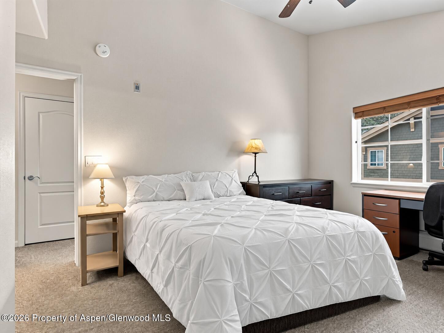 Bedroom with a large white bed, dark dresser, and desk beside a window.