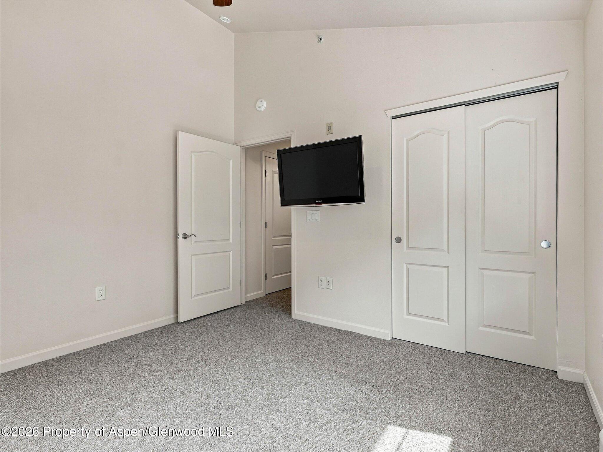 Empty bedroom with gray carpet, wall-mounted television, and white sliding closet doors.