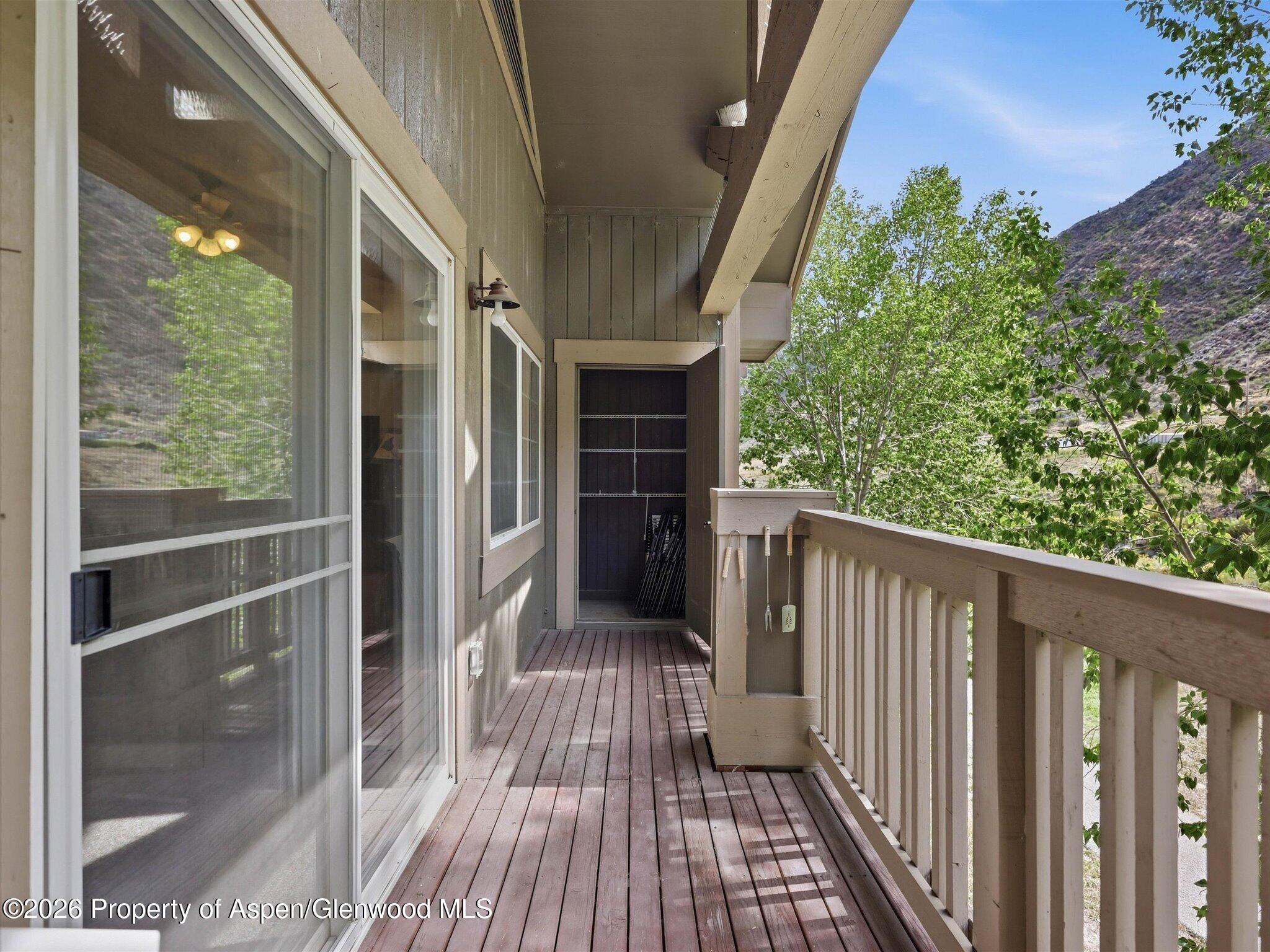 Covered wooden balcony with sliding glass doors, railing, and views of leafy trees and a rocky hillside.
