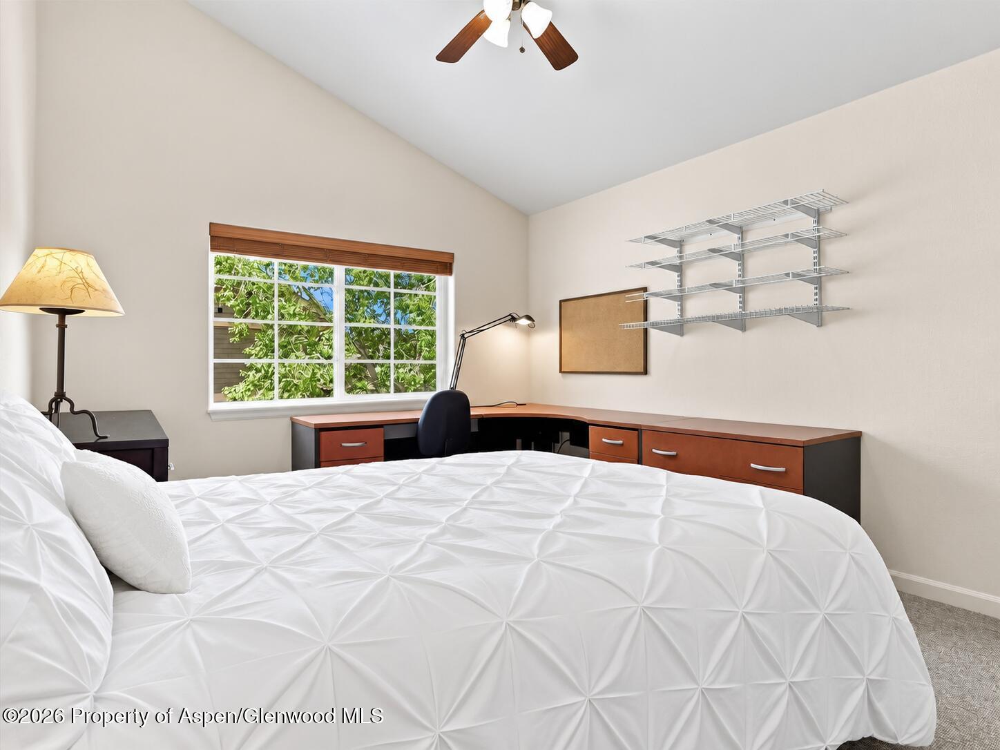 Bedroom with a large white bed, L-shaped desk, vaulted ceiling, and window overlooking leafy trees.