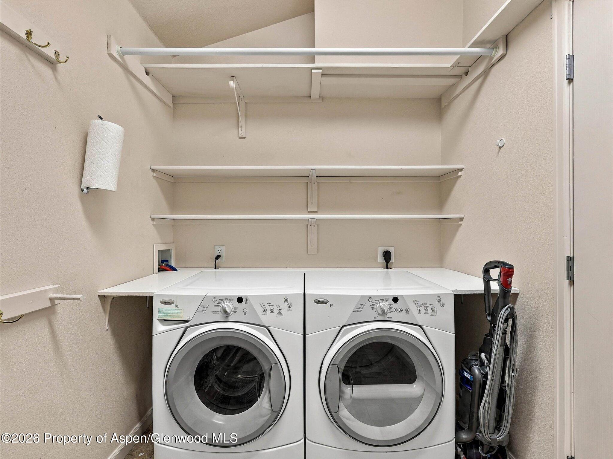Laundry room with front-loading washer and dryer, storage shelves, and a vacuum.