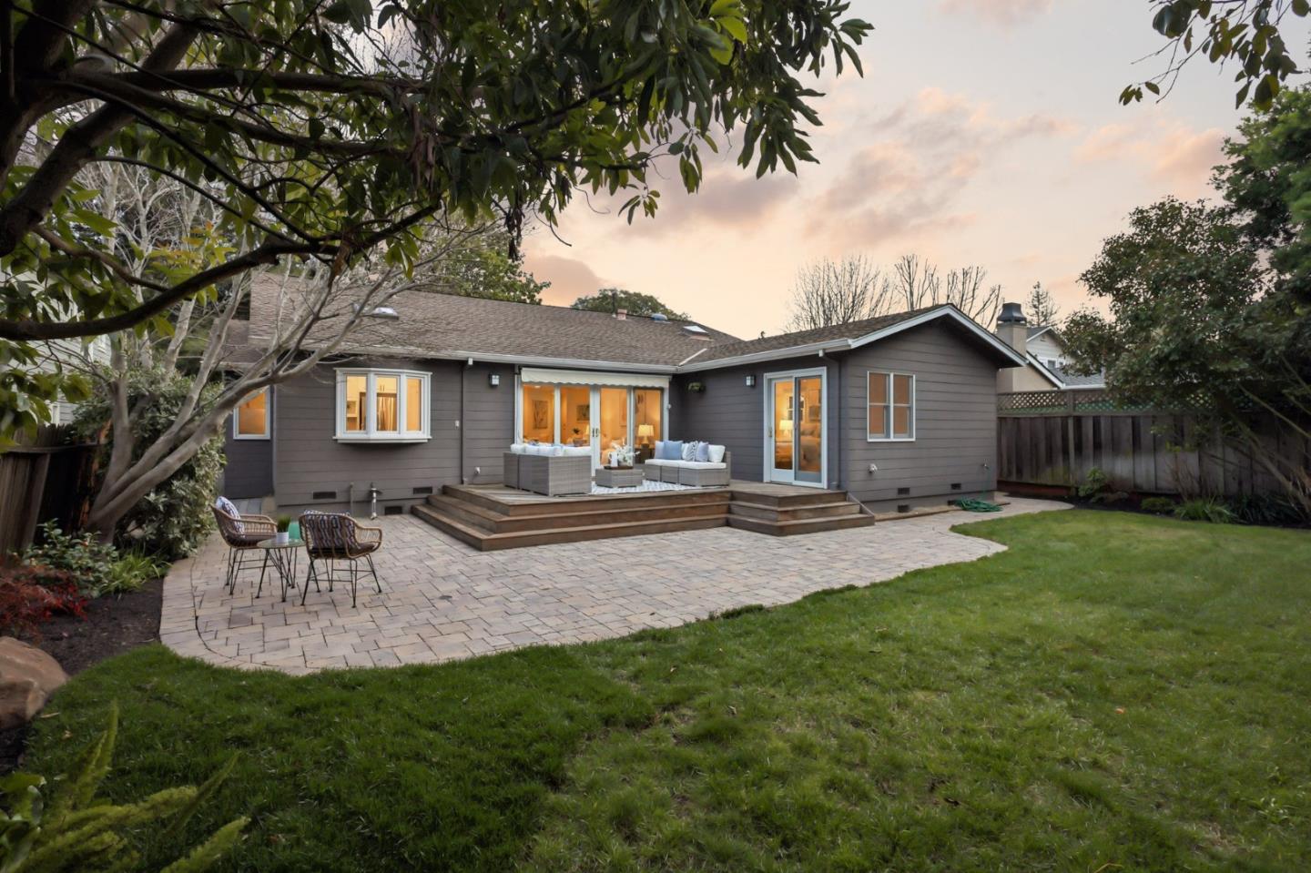 Gray single-story home with a raised deck, paved patio seating area, and lawn at sunset.