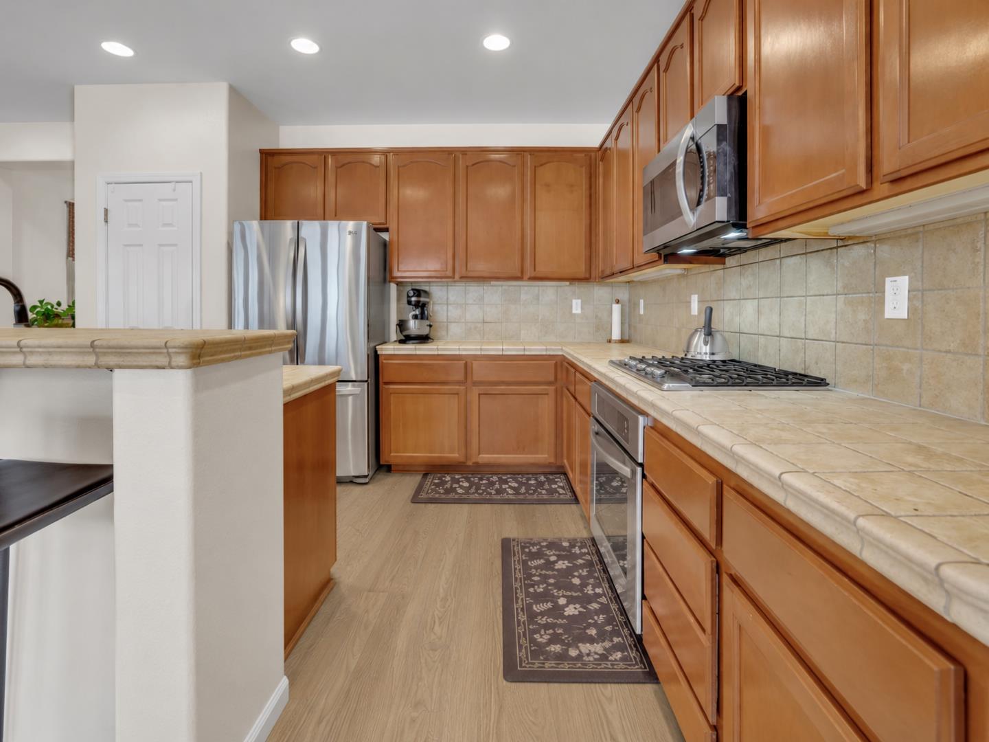 Kitchen with warm wood cabinetry, tiled counters, stainless steel appliances, and patterned floor runners.