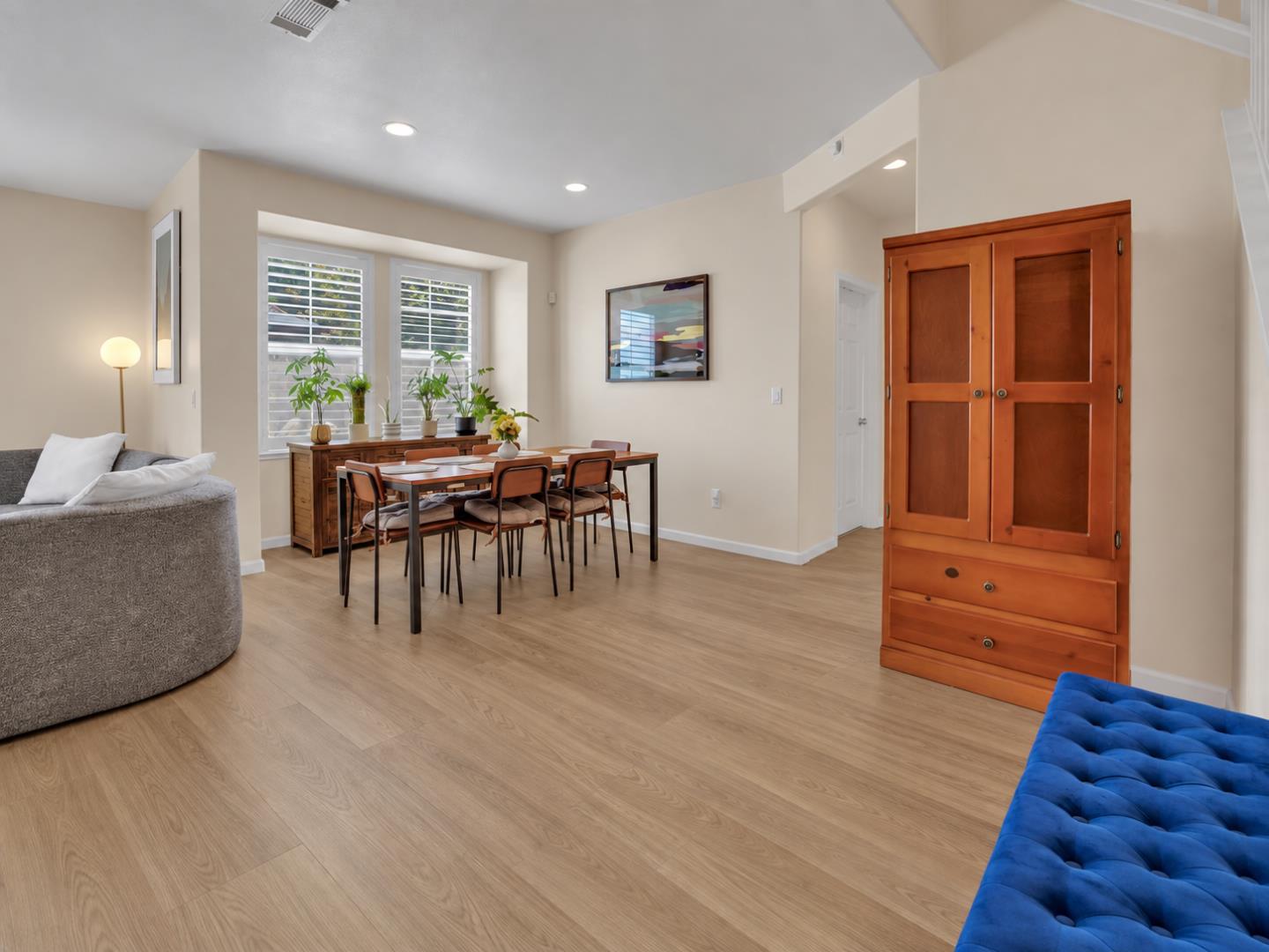 Open-plan dining and living area with shuttered windows, light wood floors, and a tall wooden cabinet.