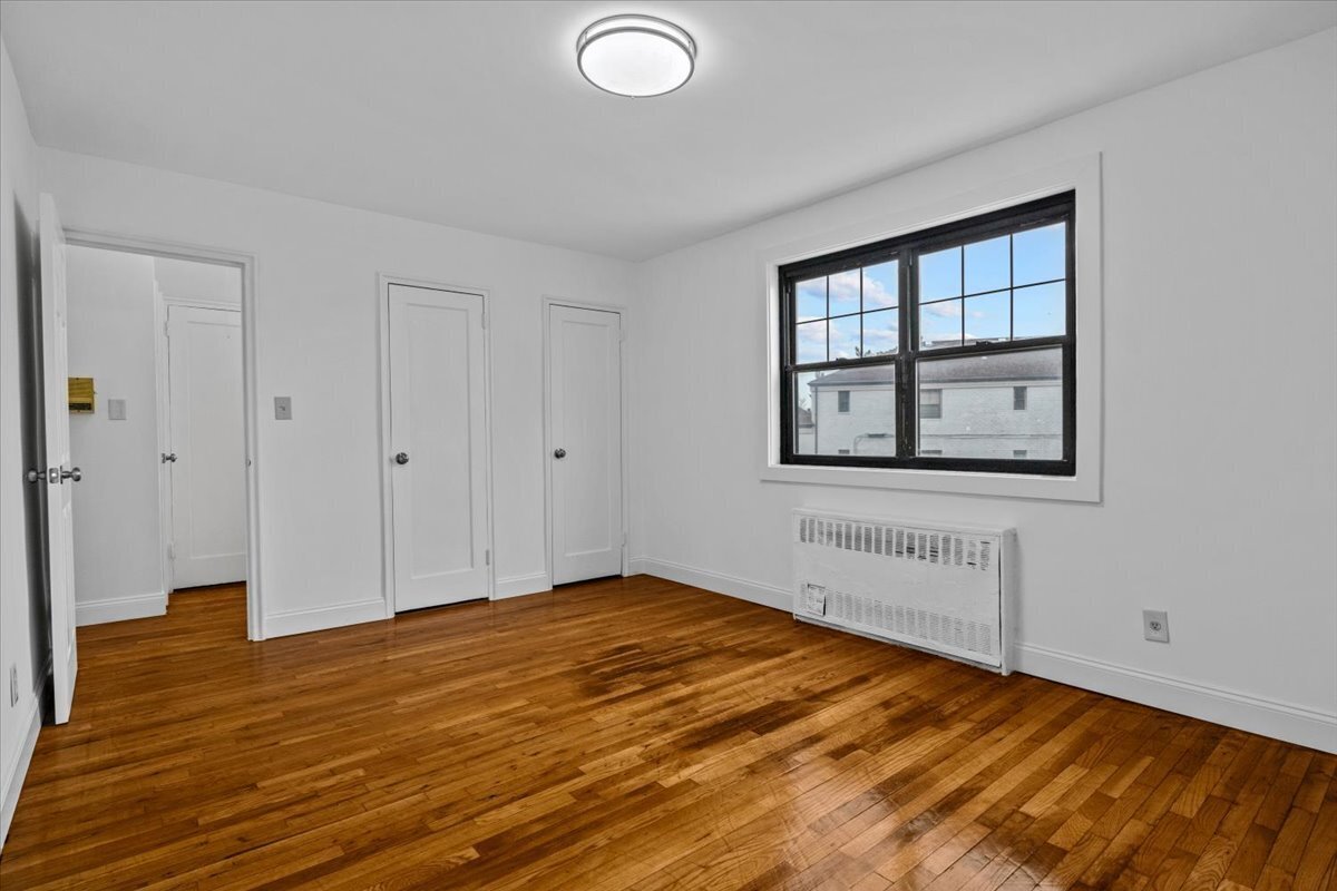 Vacant room with polished hardwood floors, three doors, and a black-framed window above a radiator.