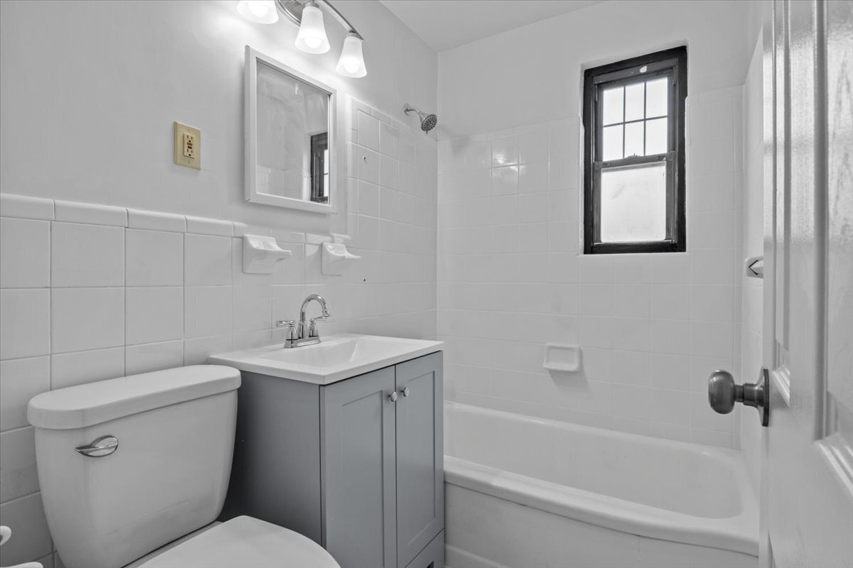 Bathroom with white tile, a bathtub and shower, gray vanity, and black-framed window.
