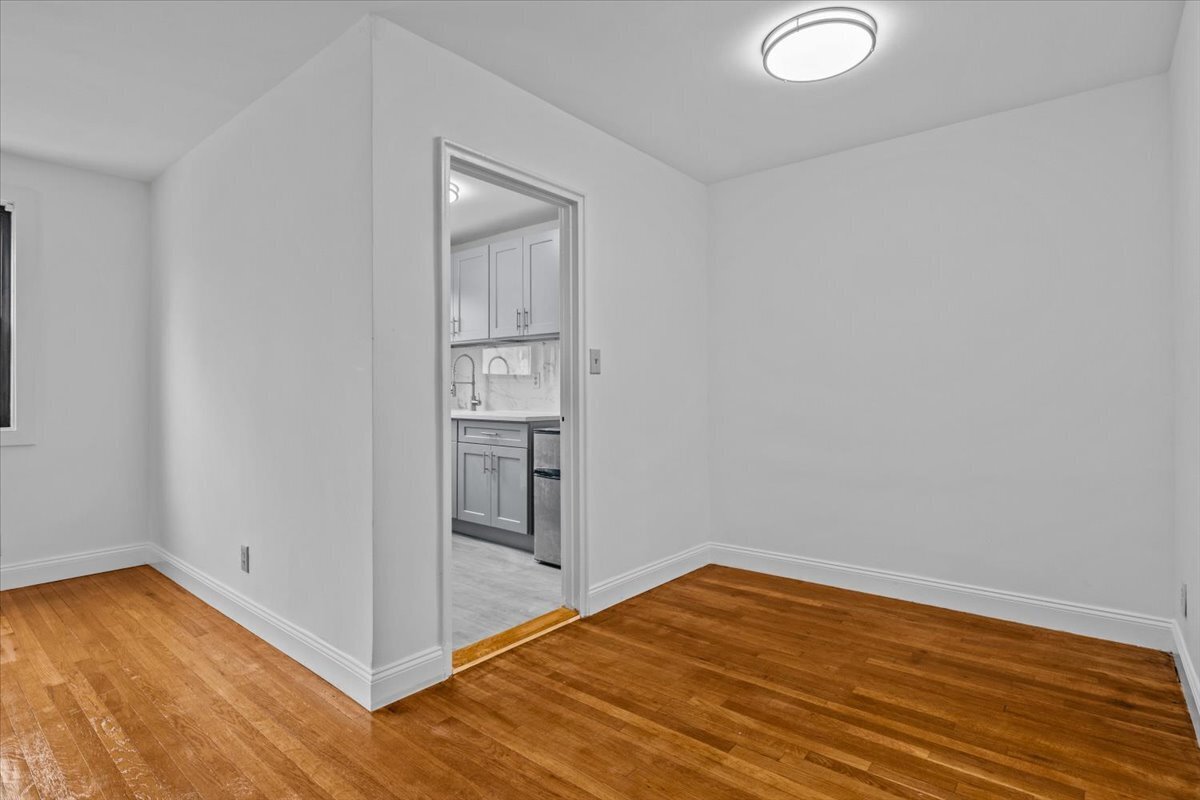Empty room with polished hardwood floors and a doorway into a gray-and-white kitchen.