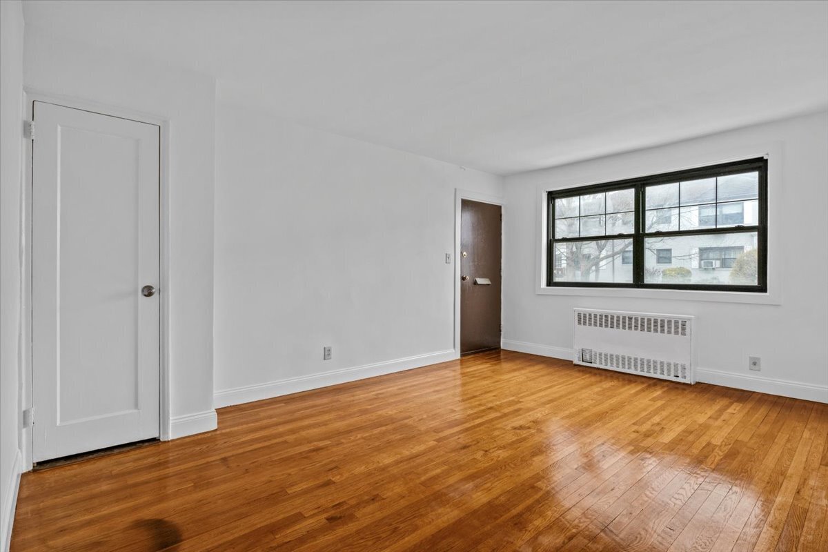 Empty apartment room with polished hardwood floors, a large window, and a radiator.