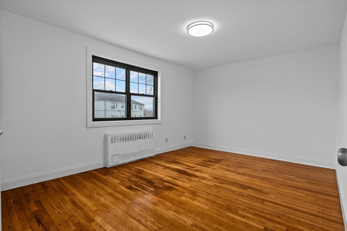 Empty room with polished hardwood floors, a black-framed window, and a wall-mounted radiator.