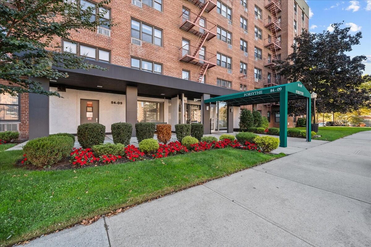 Red-brick apartment building entrance with a green Forsythe canopy and landscaped flower beds.