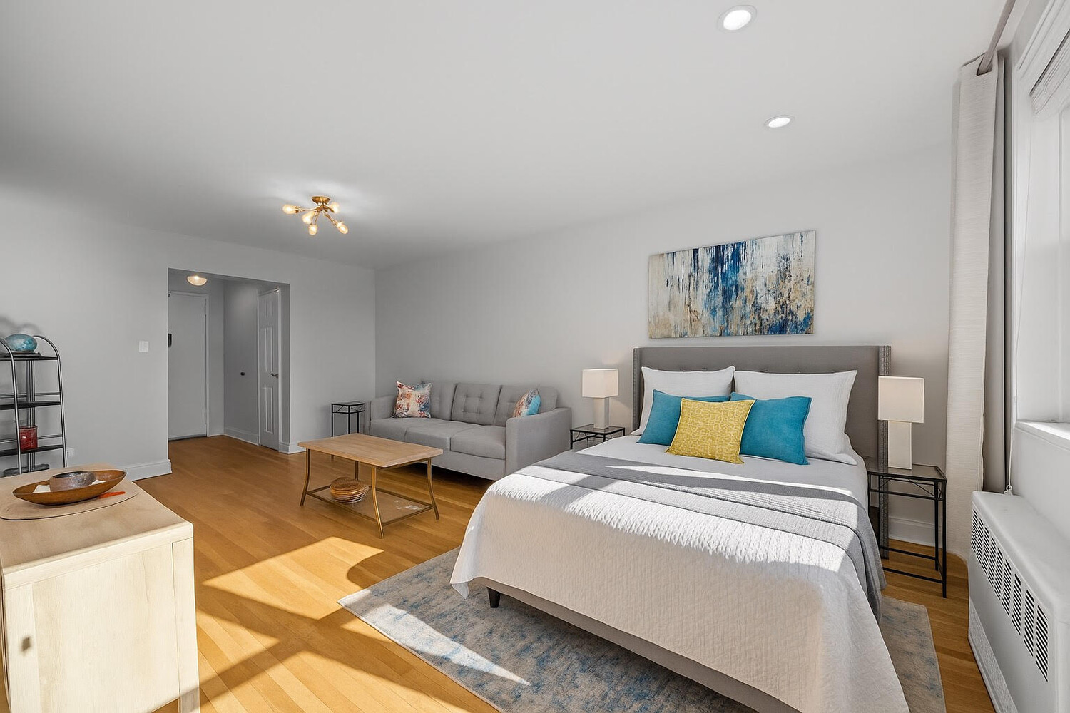 Staged bedroom with a large bed, colorful accent pillows, and a gray sofa beside a sunlit window.