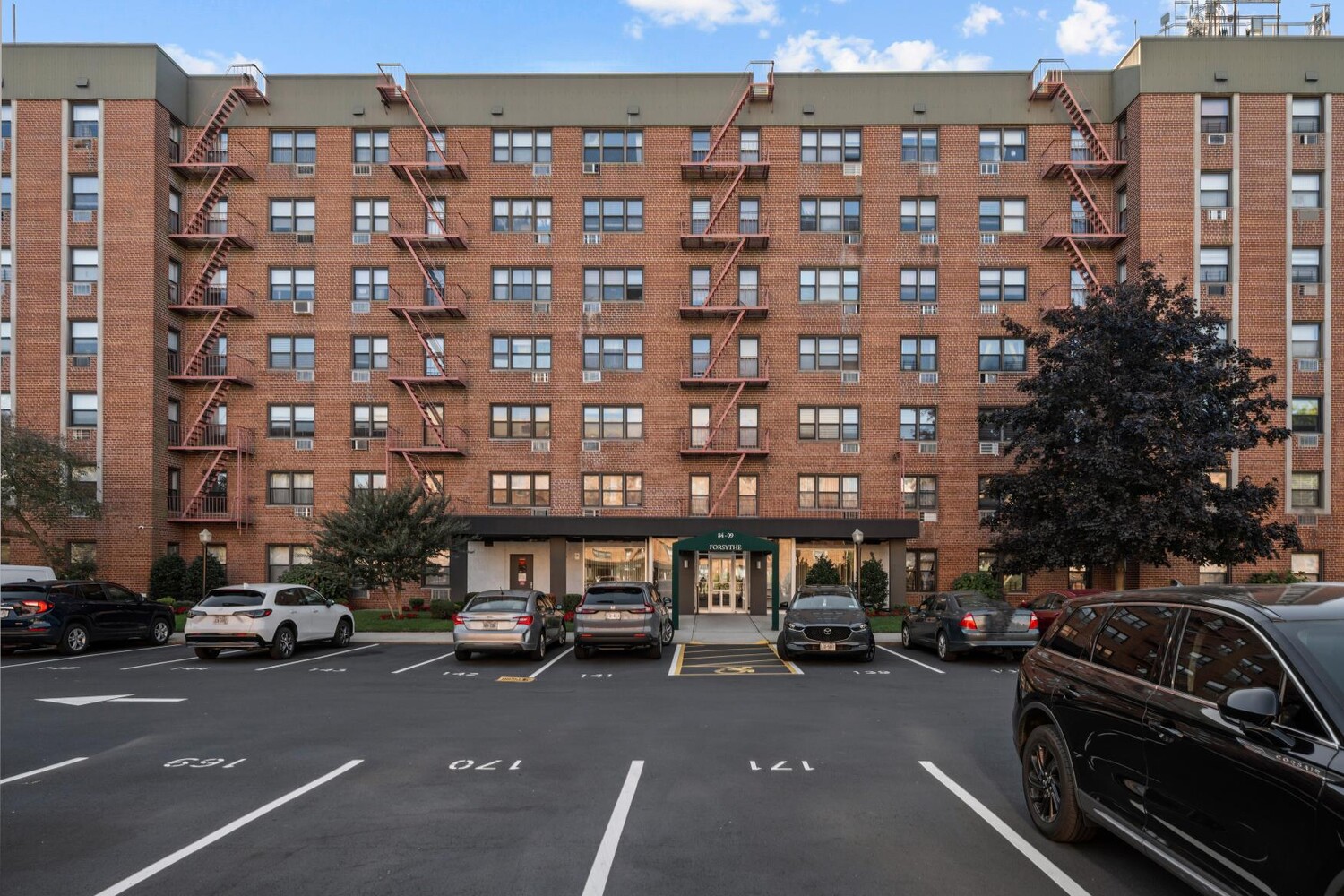 Red-brick apartment building with exterior fire escapes and a central entrance facing a parking lot.