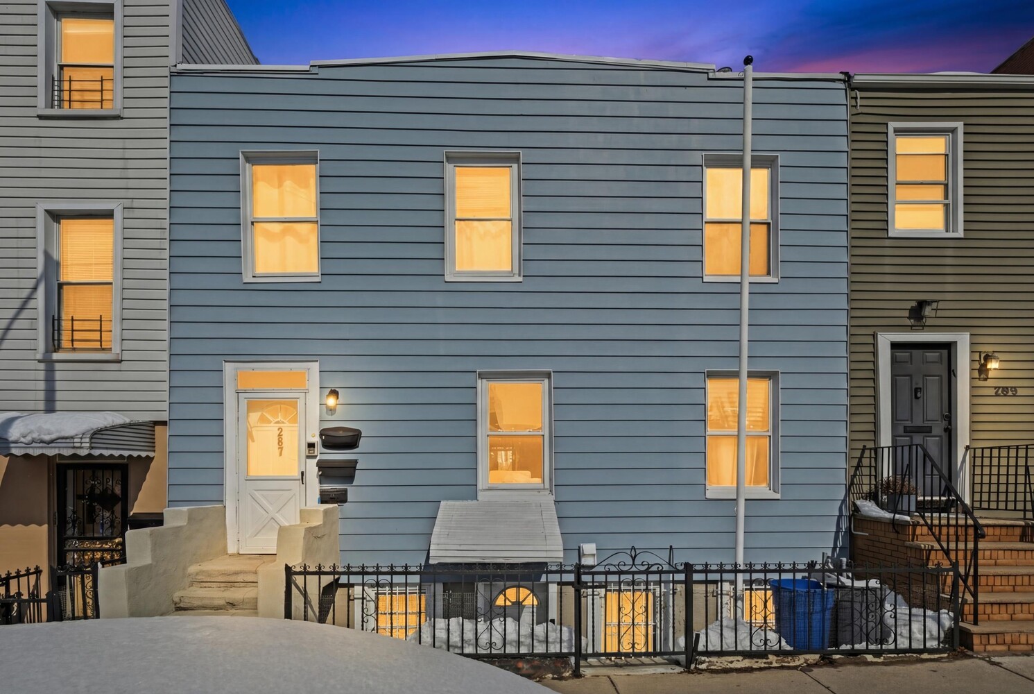Blue two-story home at dusk, with warmly lit windows and snow along the sidewalk.