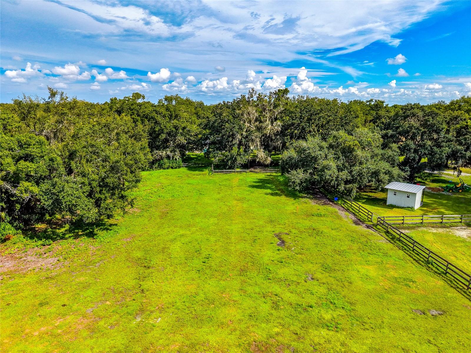 Aerial view of a grassy property with mature trees, fenced sections, and a small white shed.