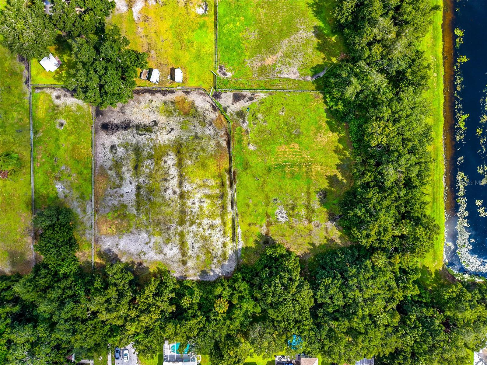 Aerial view of fenced grassy land beside a narrow waterway, surrounded by trees.