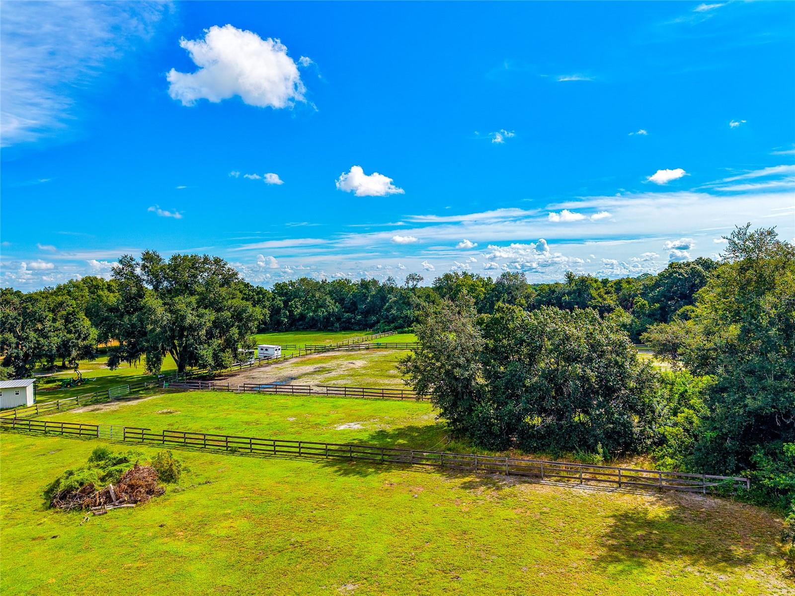Aerial view of fenced green pasture bordered by mature trees, with a small outbuilding.