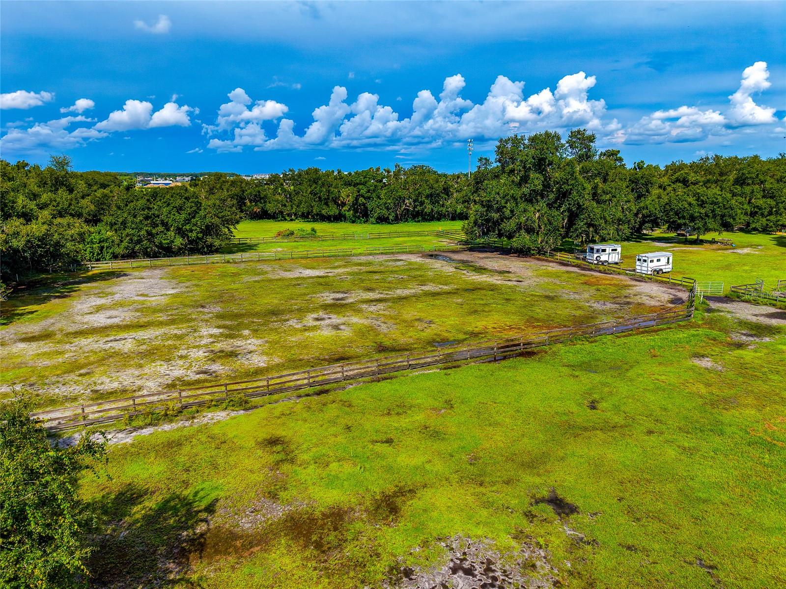 Aerial view of fenced grassy paddocks with two trailers and a surrounding tree line.