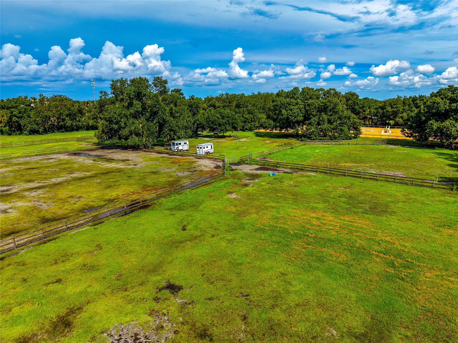 Aerial view of fenced green pastures with mature trees, two trailers, and a riding arena.