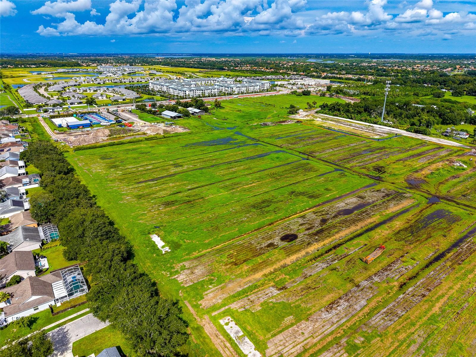 Aerial view of a vacant grassy parcel with drainage channels, beside homes and new construction.