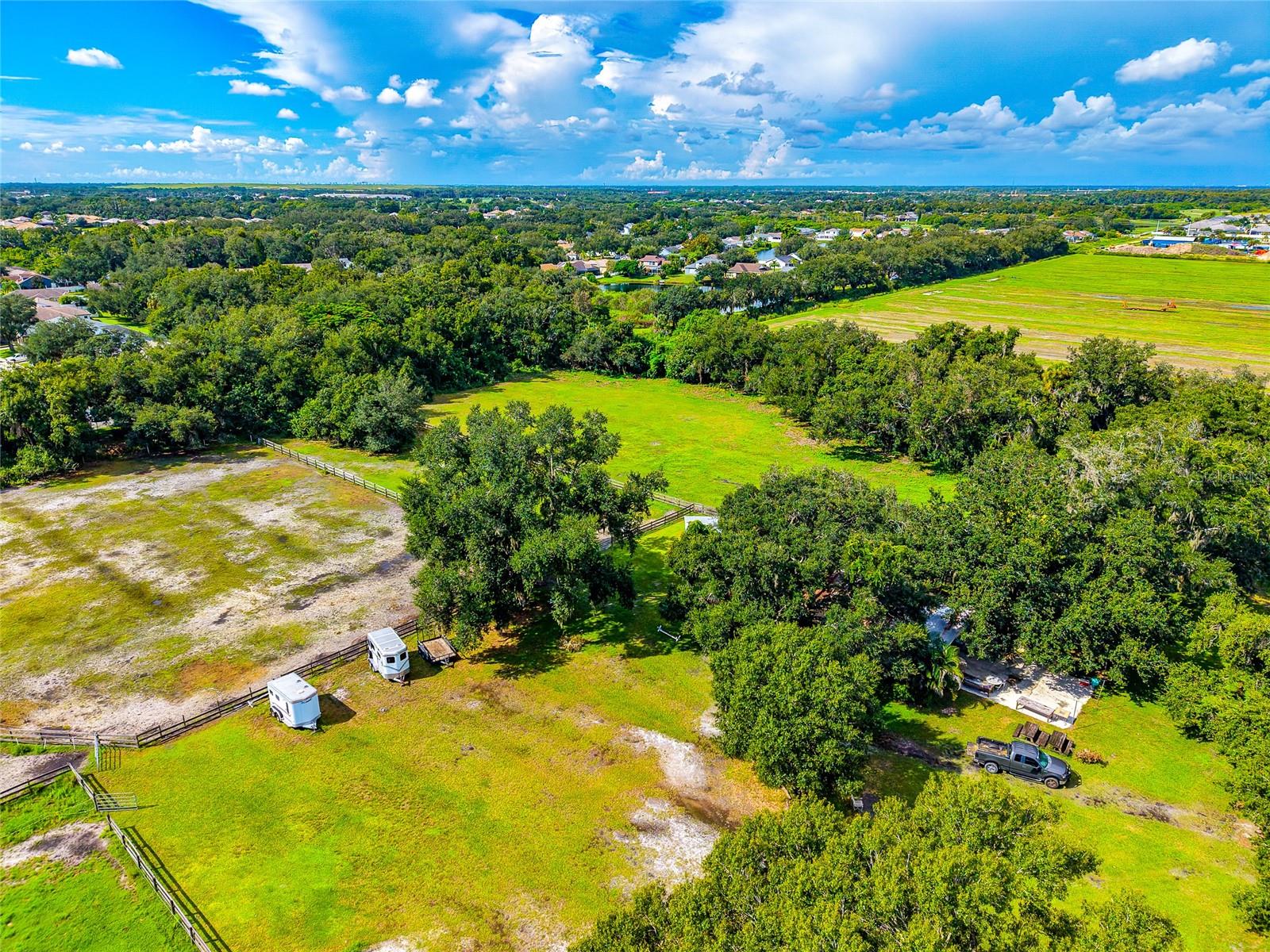Aerial view of vacant grassy land with fenced areas, mature trees, two trailers, and a parked truck.
