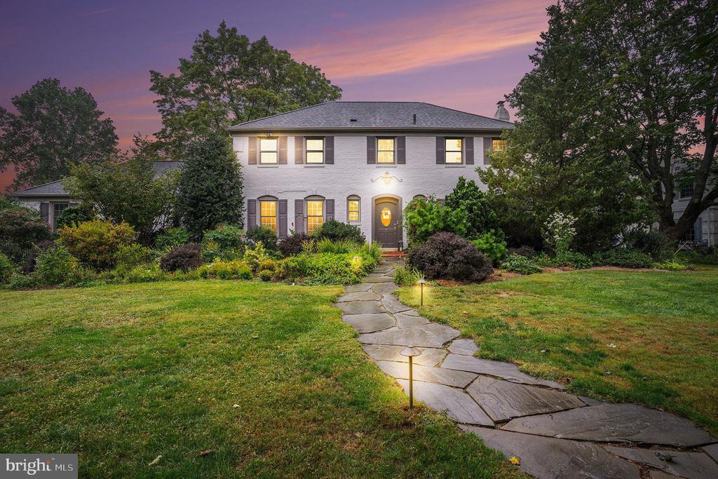 Two-story white brick house with lit windows and a stone walkway at dusk.