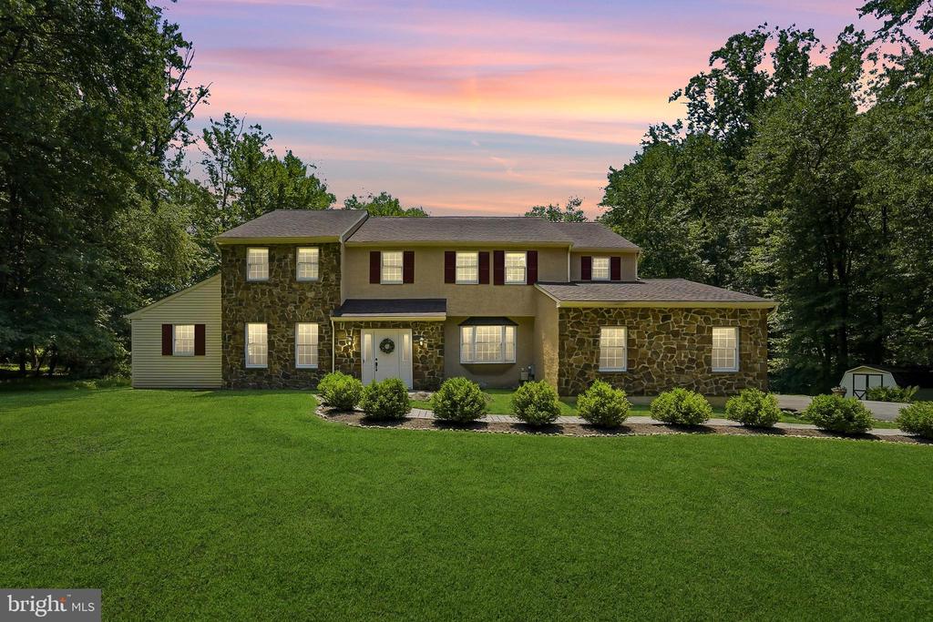 Two-story stone and beige house with a broad lawn and mature trees at dusk.