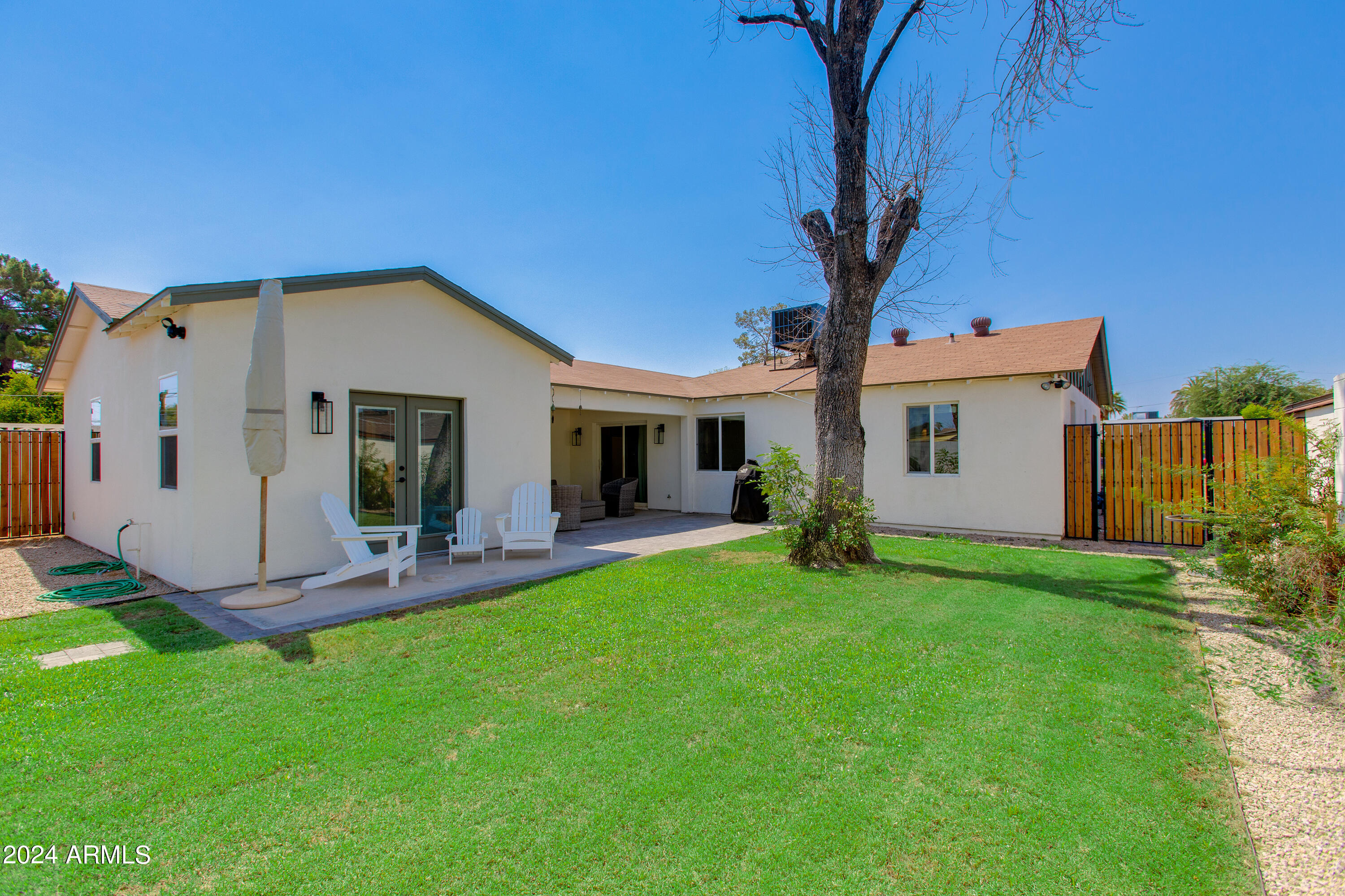 Home’s backyard with a covered patio, white chairs, large lawn, and mature tree.