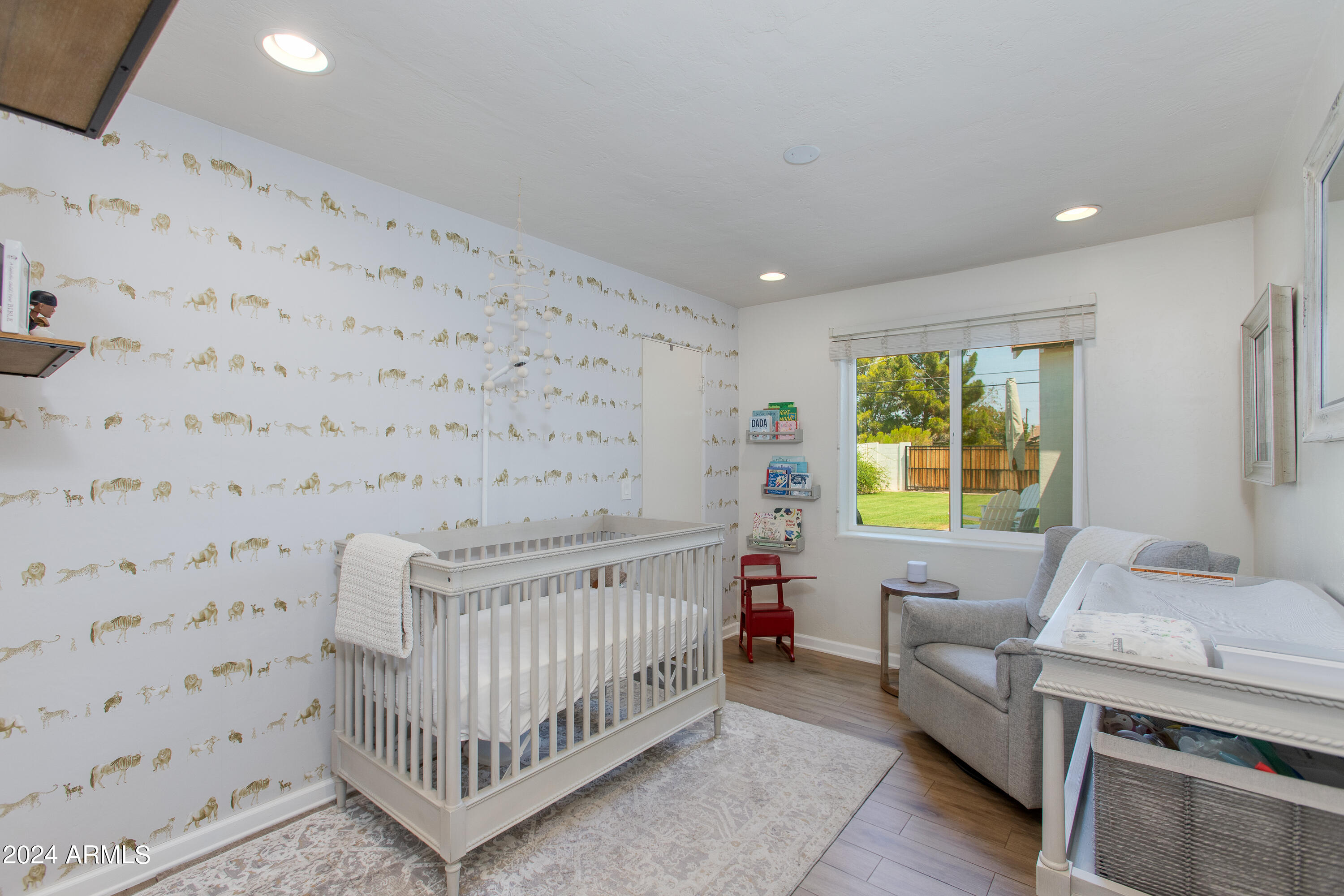 Nursery with a white crib, animal-patterned wallpaper, gray armchair, and window overlooking the yard.