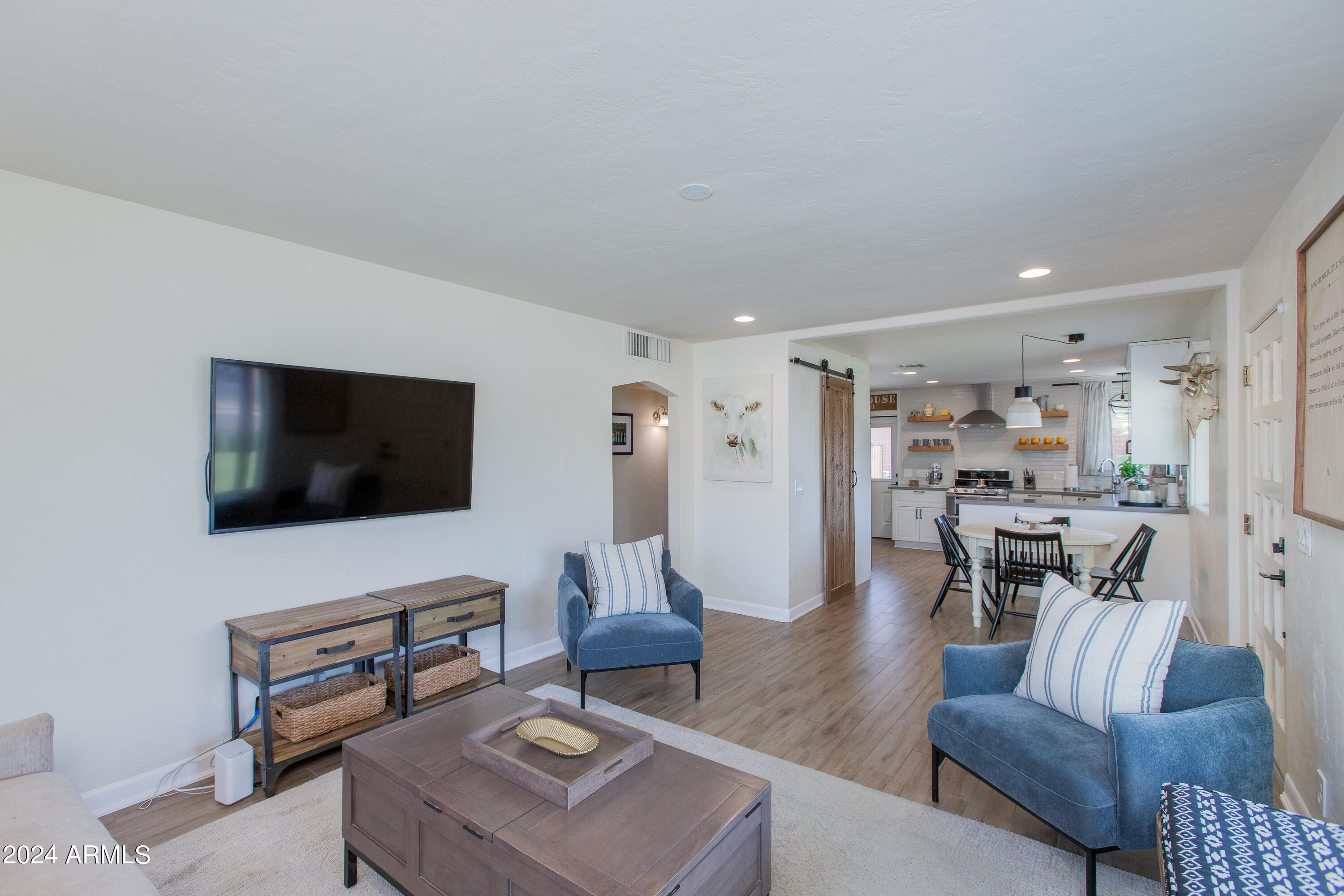Staged living room with blue chairs, wall-mounted TV, and an open view into the dining area and kitchen.