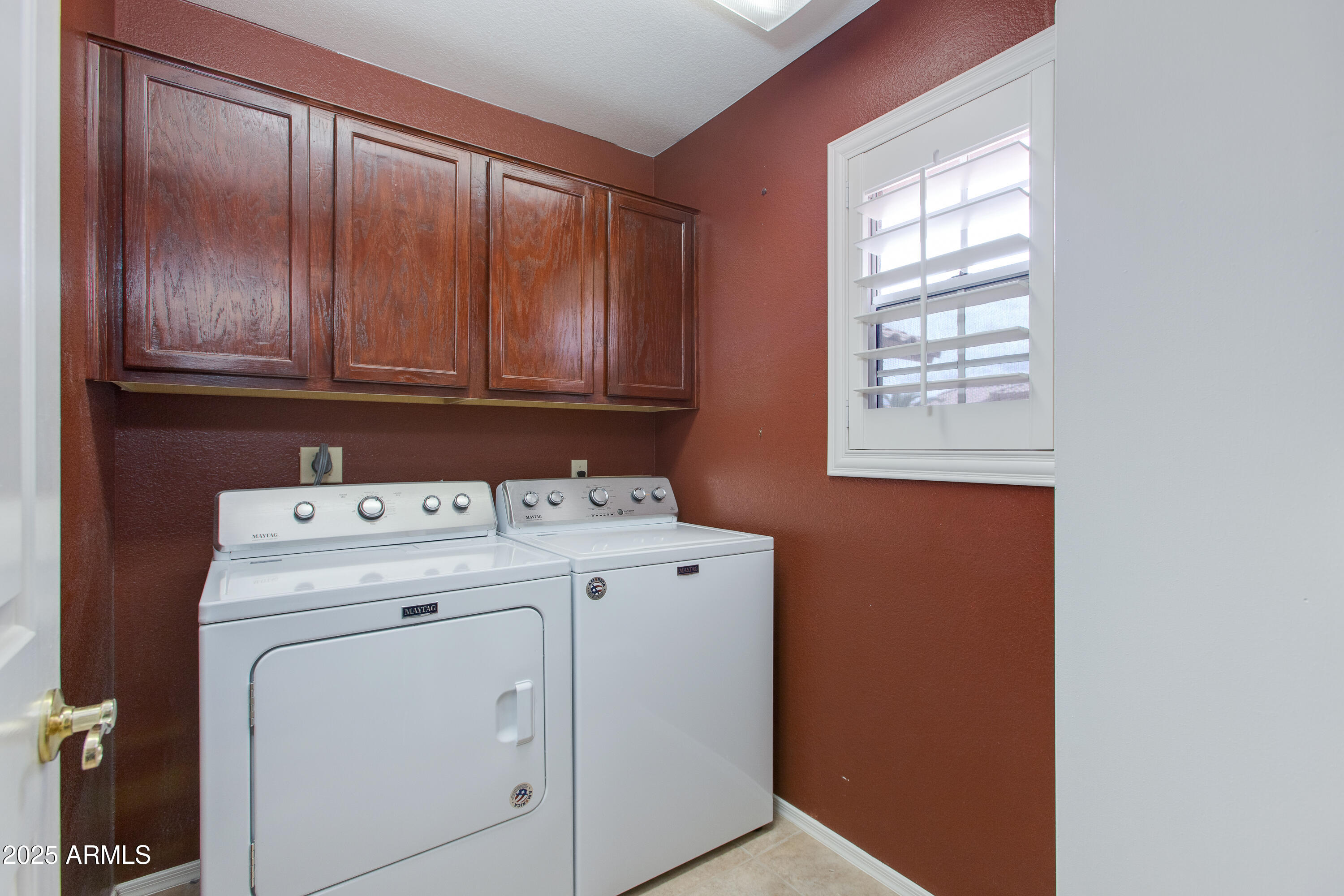 Laundry room with a Maytag washer and dryer beneath dark wood cabinets and beside a shuttered window.