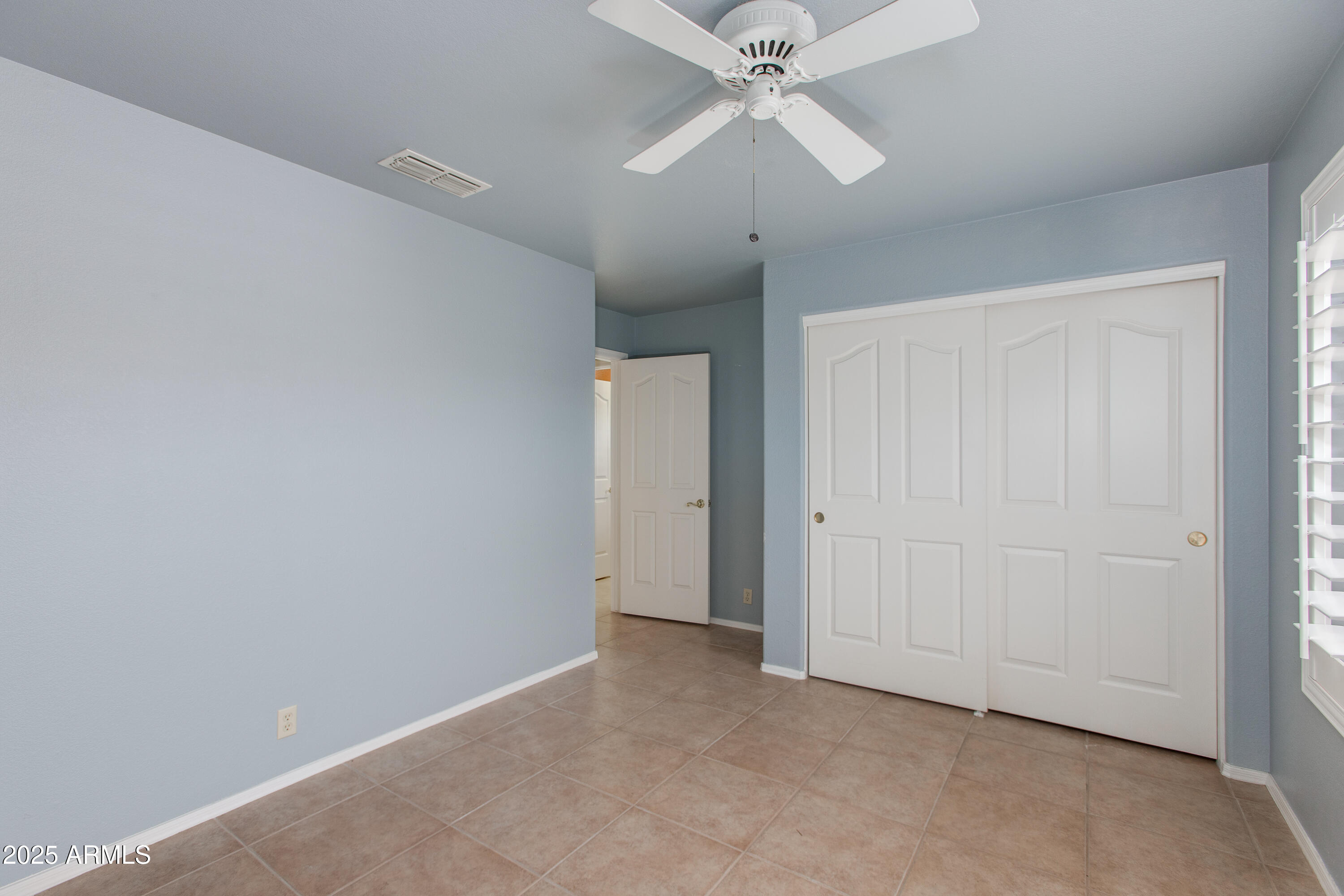 Empty bedroom with blue walls, tiled floor, ceiling fan, and white sliding closet doors.