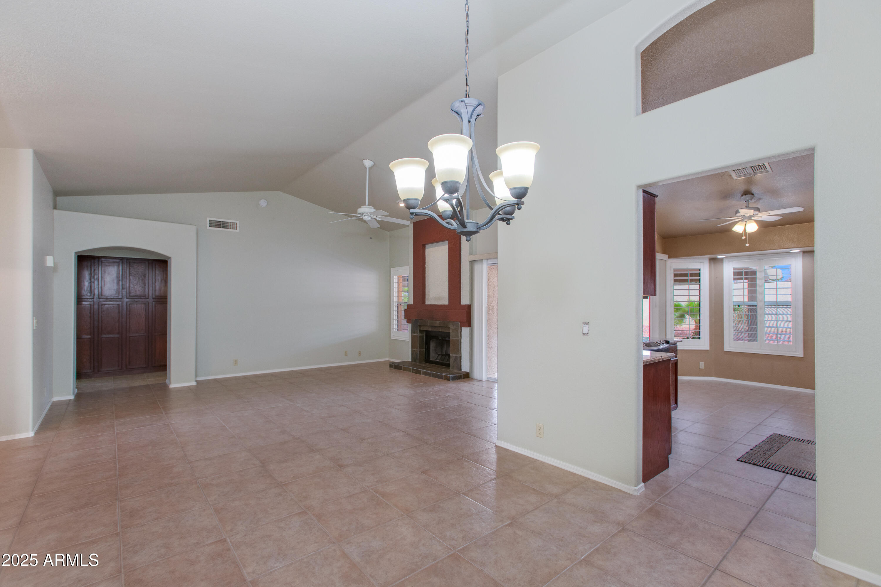 Unfurnished tiled living and dining area with a fireplace, chandelier, and adjoining room with shuttered windows.