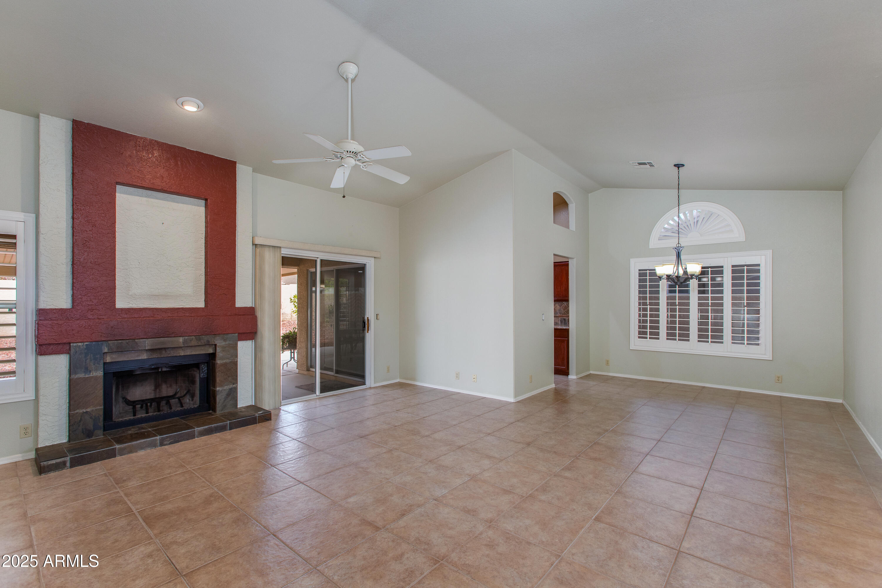Vacant living room with a red-surround fireplace, tiled floor, ceiling fan, and shuttered window.