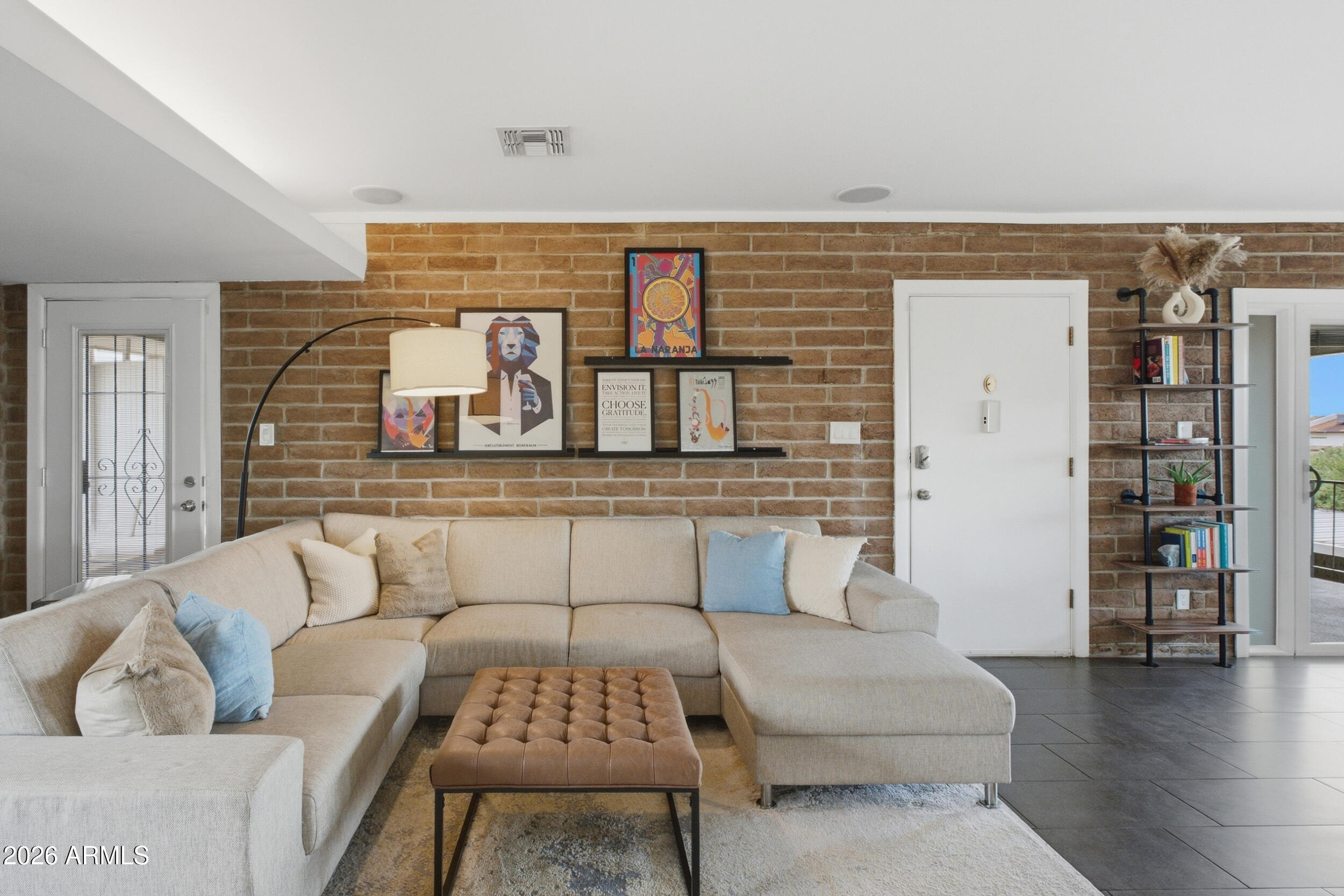Furnished living room with a beige sectional, tufted ottoman, and exposed brick wall.