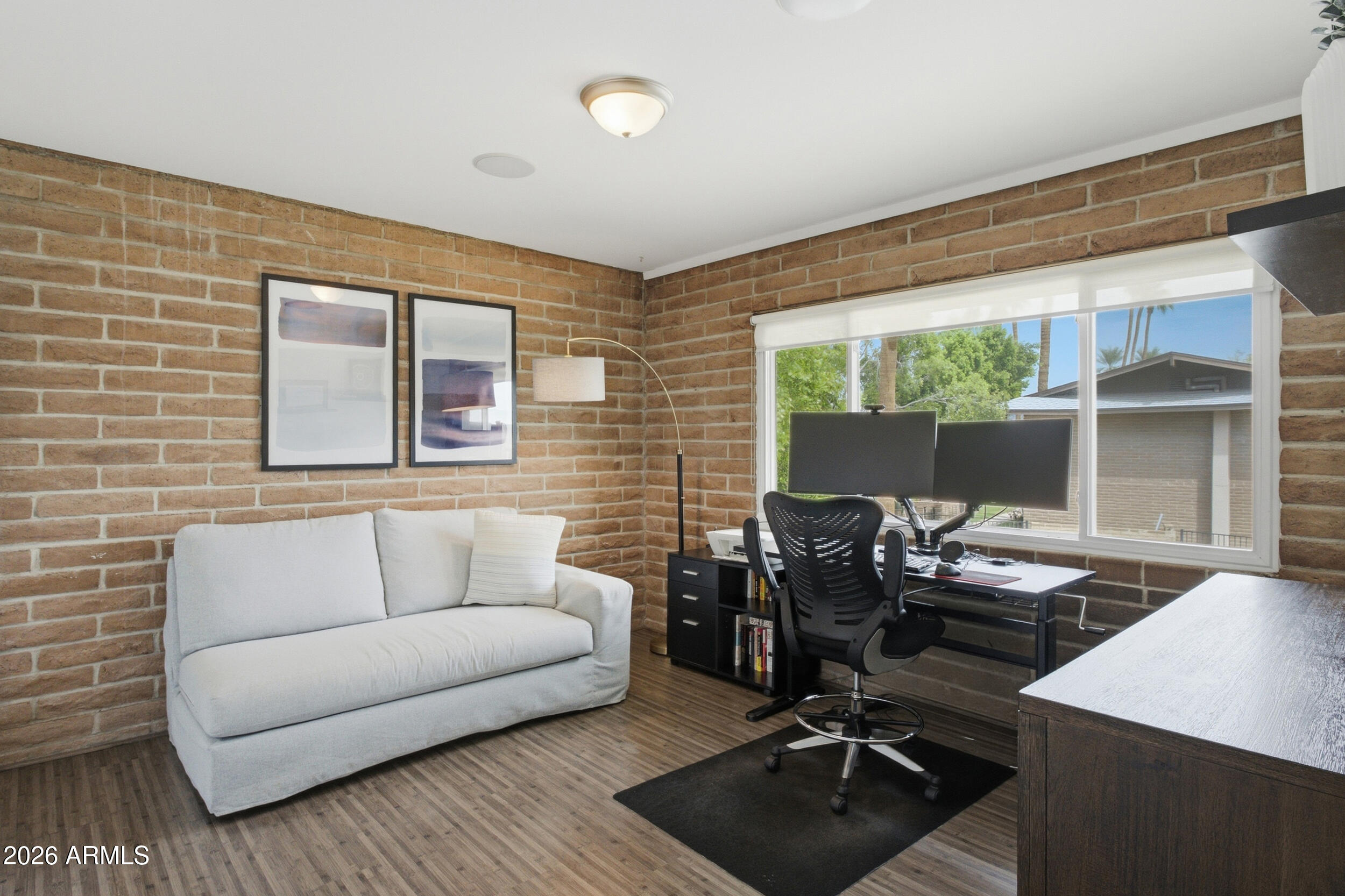Home office with exposed brick walls, a white sofa, and a desk with dual monitors beside a large window.