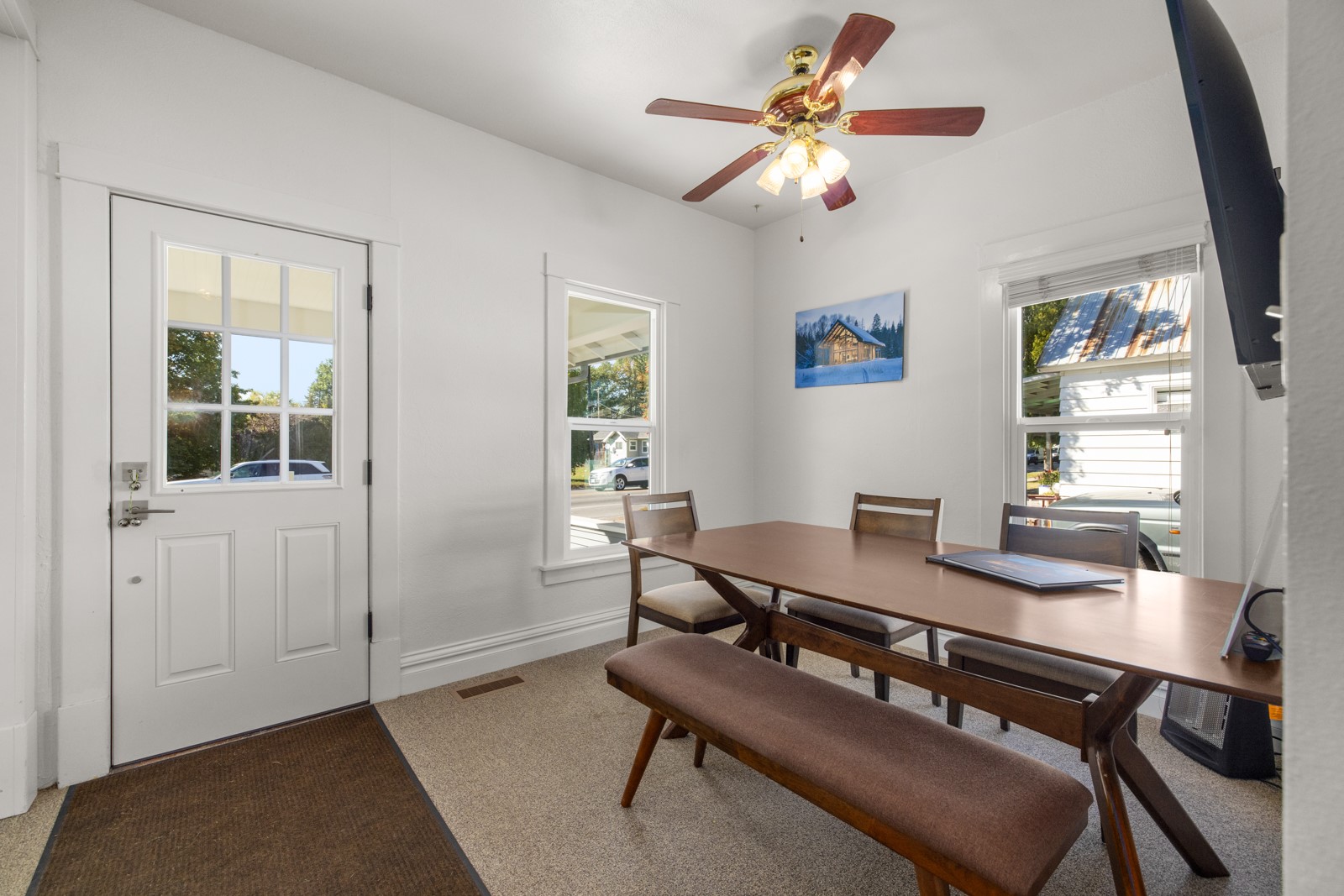 Dining area with a long wooden table, upholstered bench, ceiling fan, and windows beside the front door.