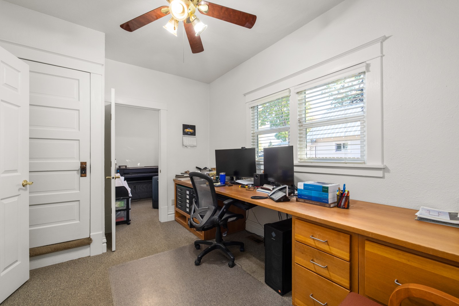 Home office with a long wooden desk, dual monitors, and windows with blinds.