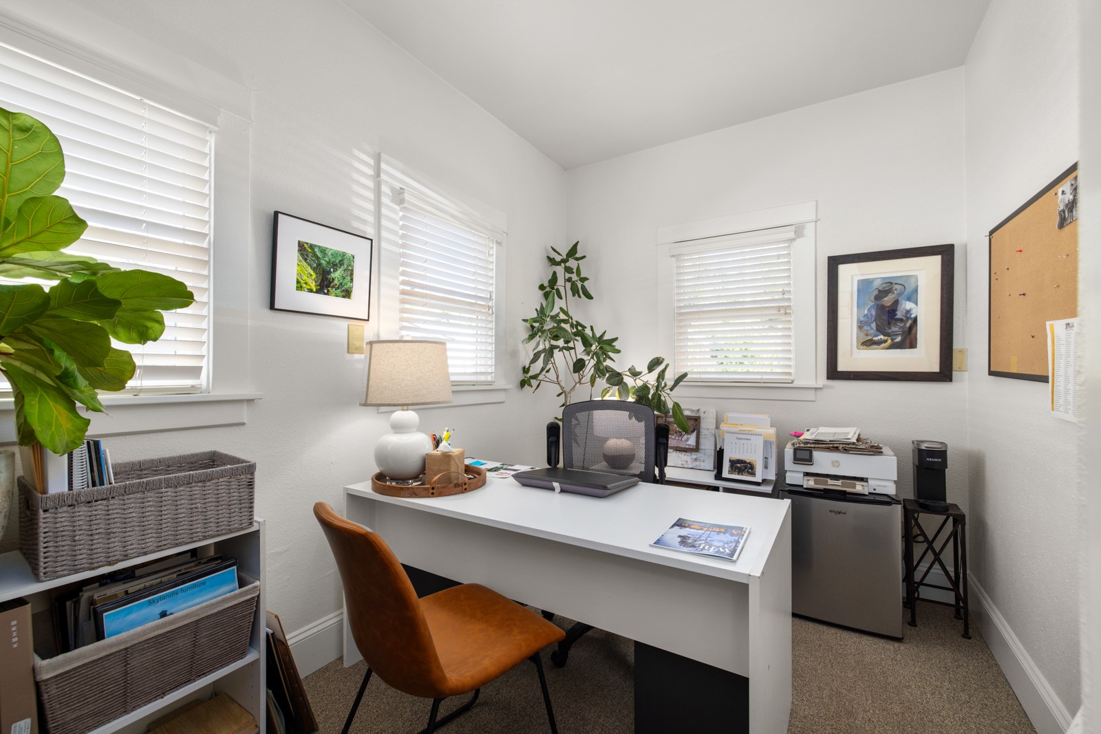 Home office with a white desk, brown chair, large windows, and leafy plants.