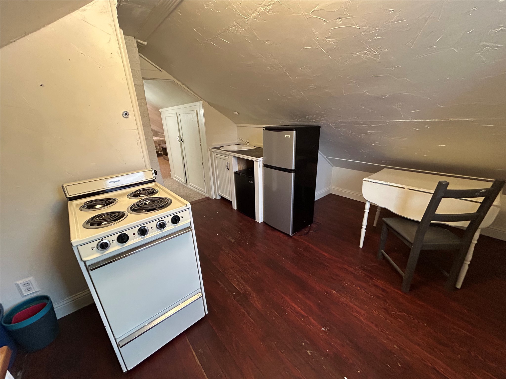 Compact attic kitchenette with an electric stove, refrigerator, sink, and small table beneath a sloped ceiling.