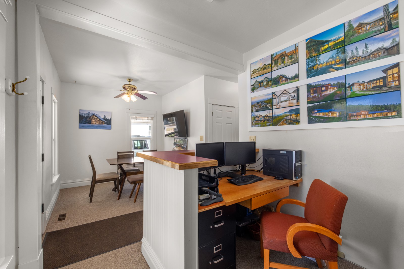 Office with a wooden desk and dual monitors, looking through to a small dining area.
