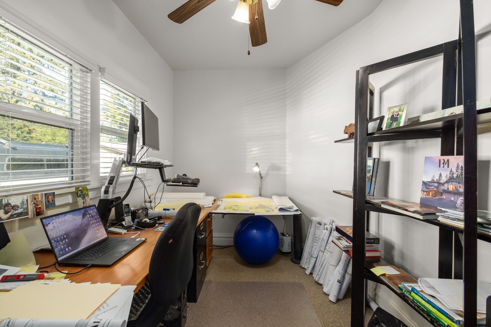 Home office with a crowded desk, black shelving, and a blue exercise ball.