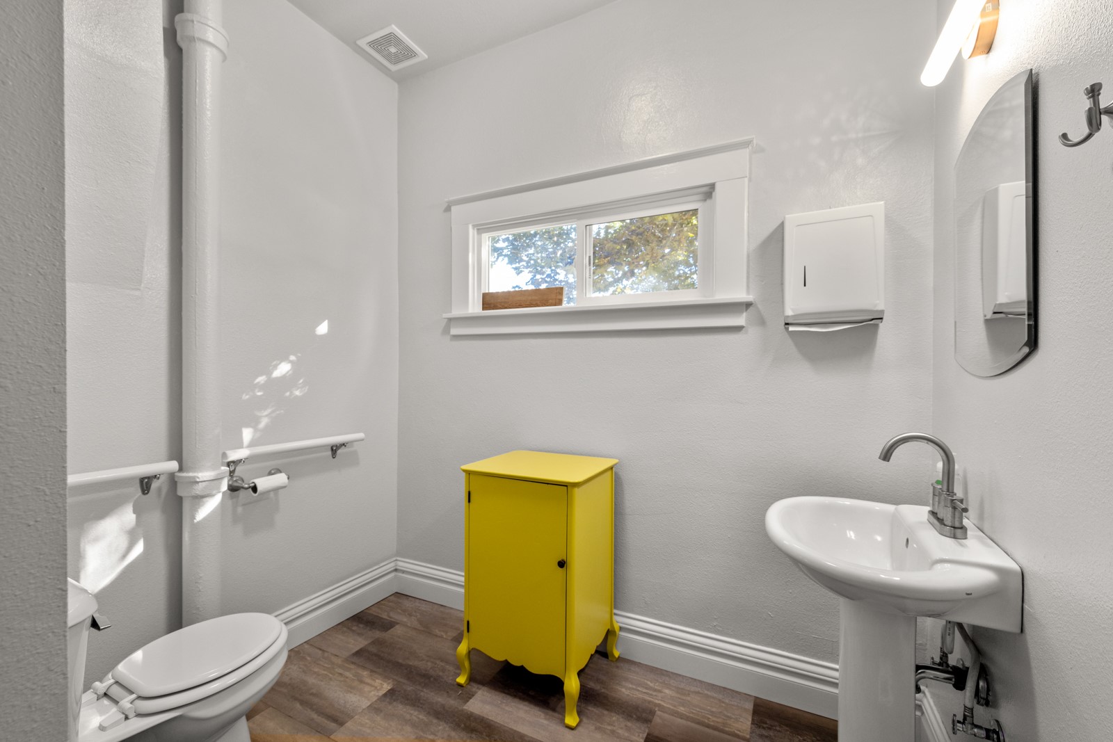 Bathroom with a pedestal sink, toilet, and bright yellow cabinet beneath a high window.
