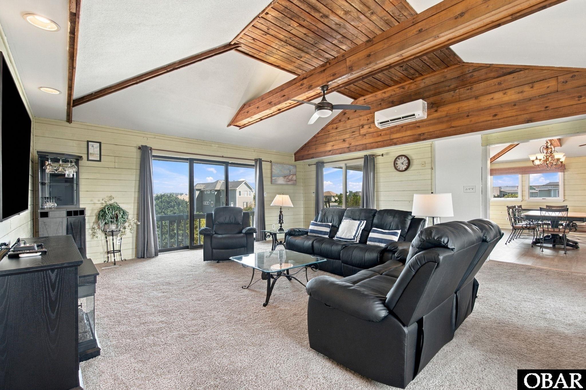 Living room with black leather seating and a vaulted wood-plank ceiling, opening to a dining area.