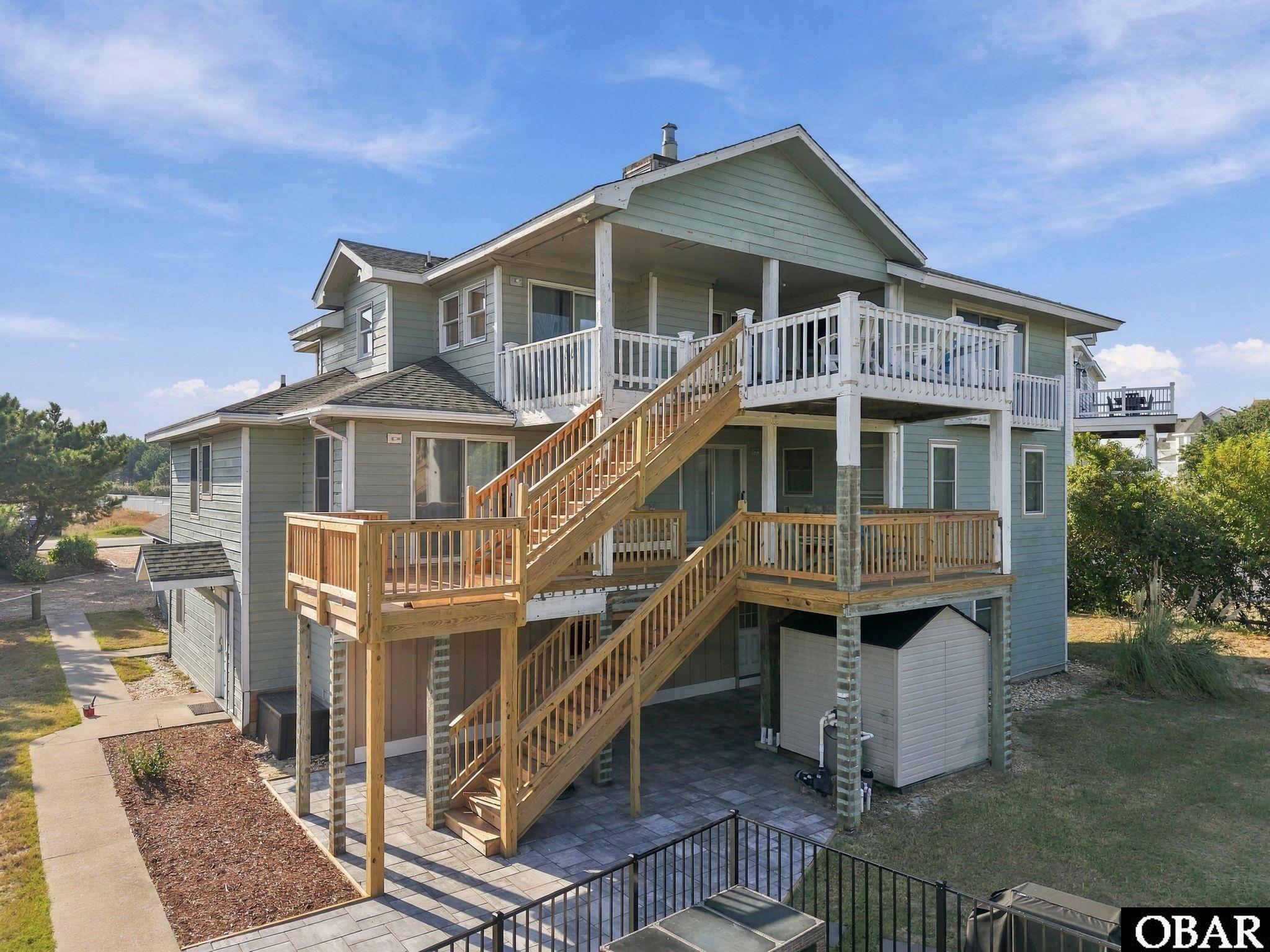 Blue-gray coastal home with spacious white decks, wooden exterior stairs, and a paved patio.