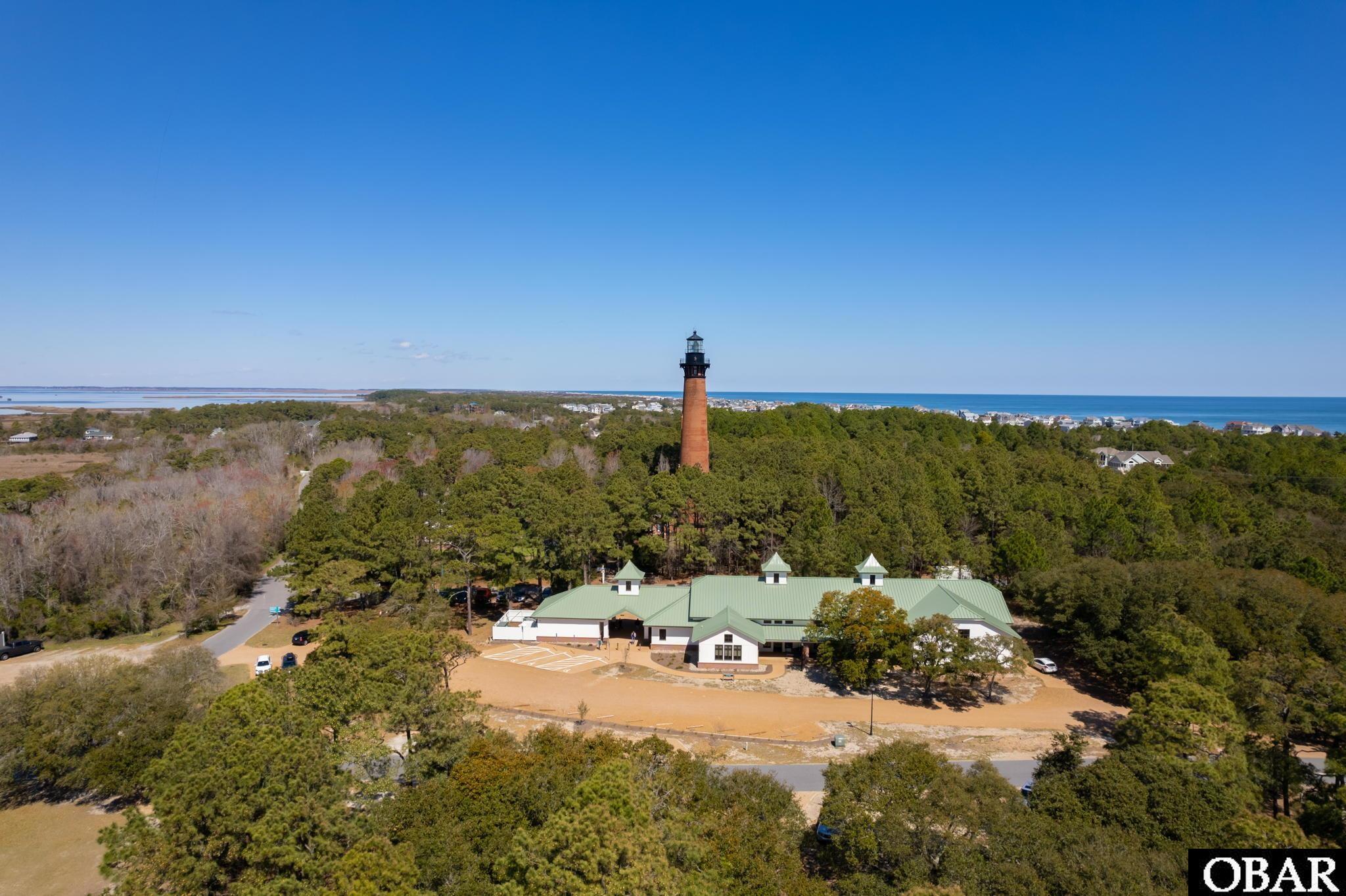 Aerial view of a brick lighthouse, green-roofed building, and ocean shoreline beyond the trees.