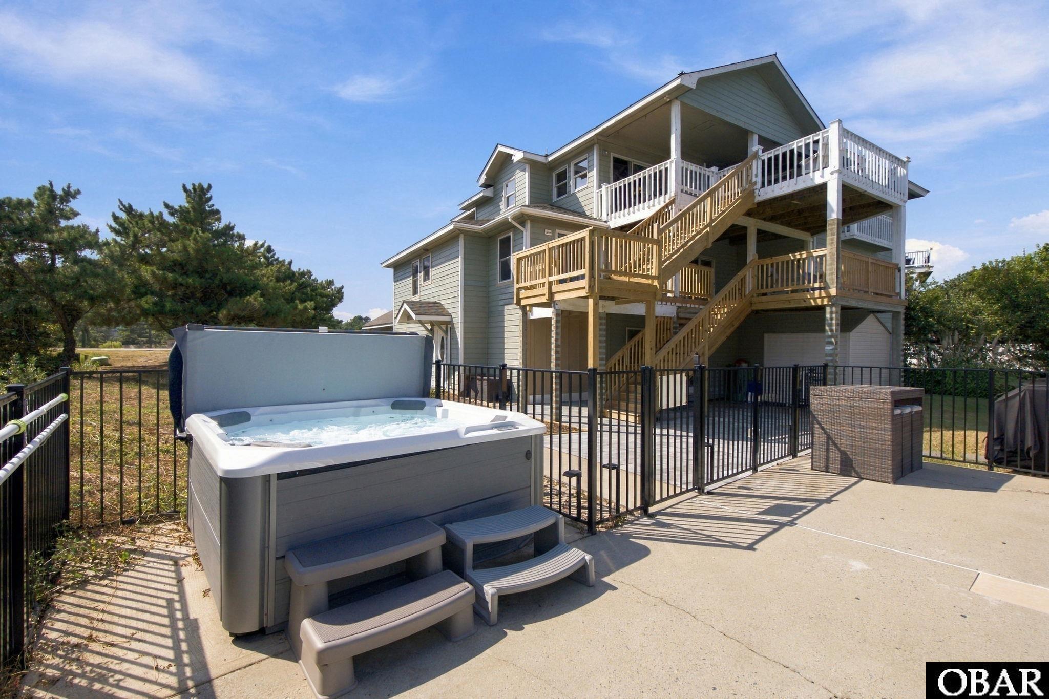 Multi-level gray home with wooden decks and stairs behind a fenced patio and hot tub.