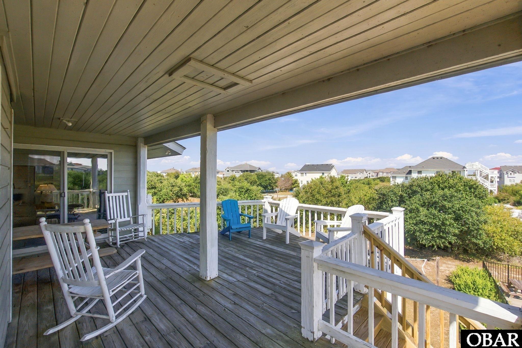 Covered wooden deck with rocking chairs and Adirondack chairs overlooking trees and nearby homes.