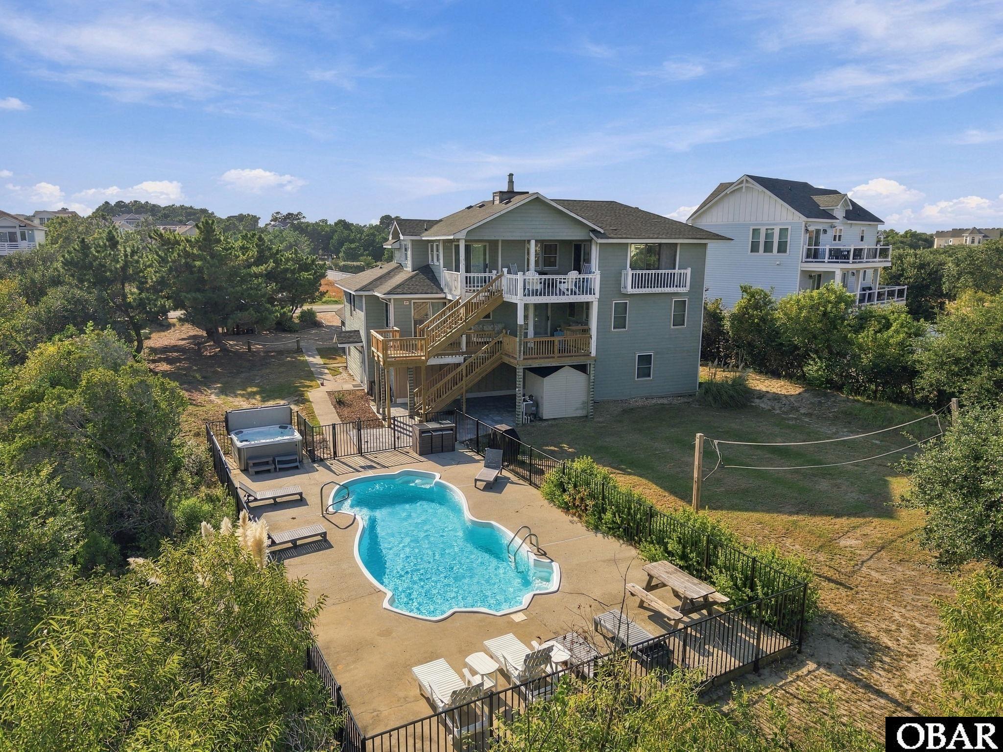 Aerial view of a gray beach house with multiple decks, a fenced pool, and hot tub.