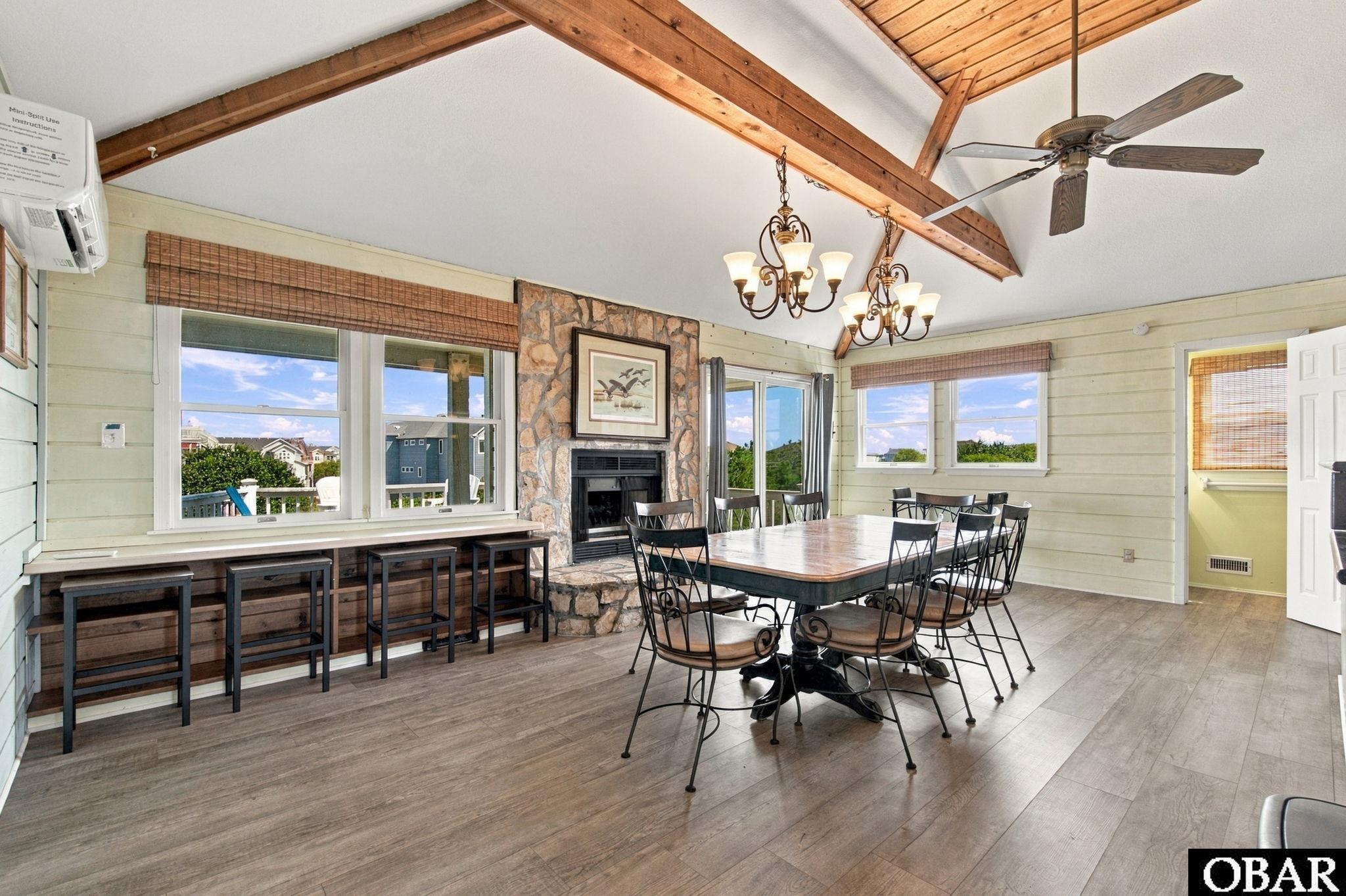 Dining area with a stone fireplace, long table, window-side counter seating, and exposed timber beams.