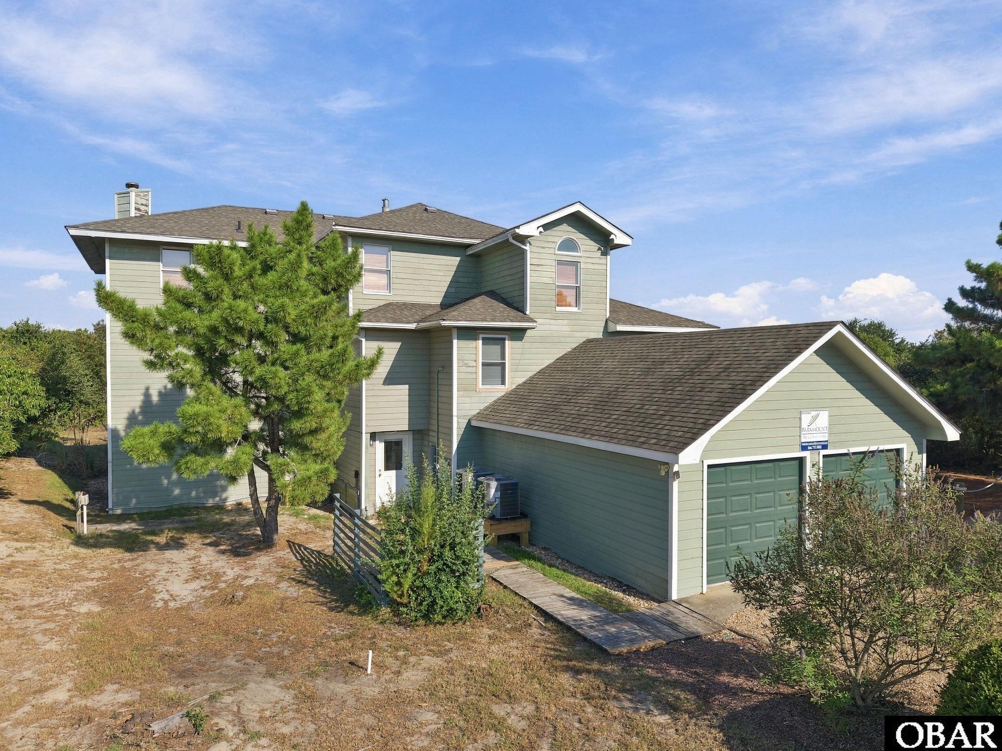 Two-story pale green house with an attached double garage and pine trees in the yard.